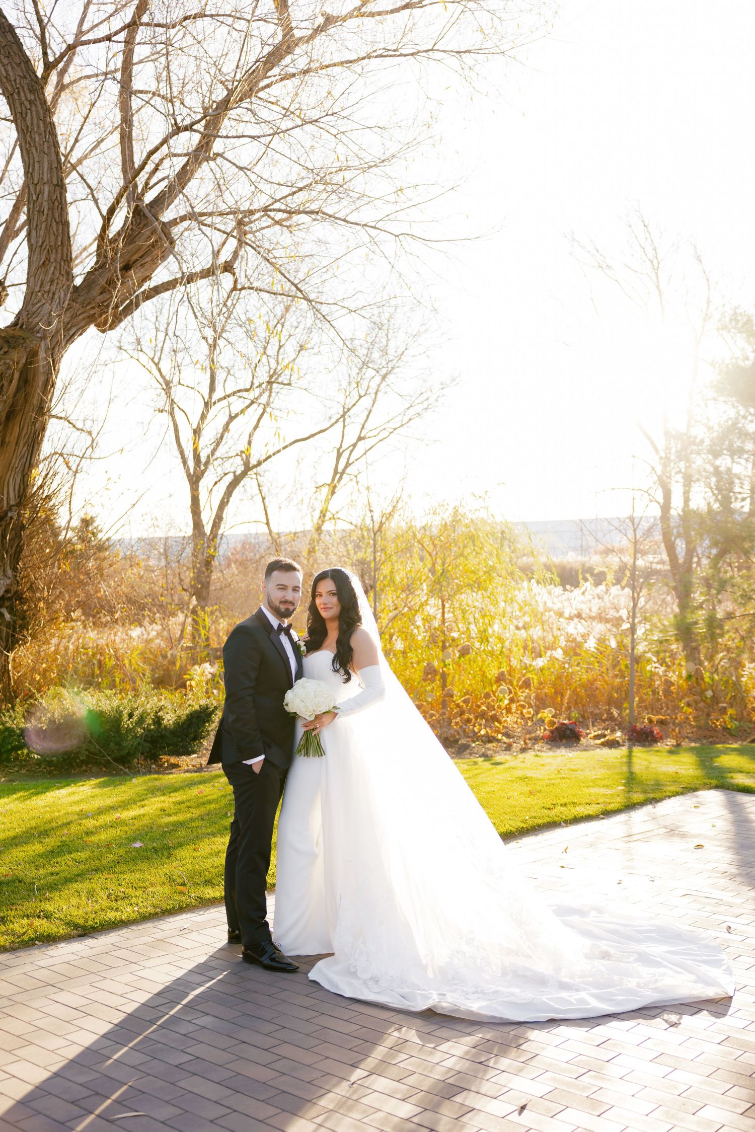A romantic wedding photo of a couple on a brick path with autumn trees and golden sunlight in the background.