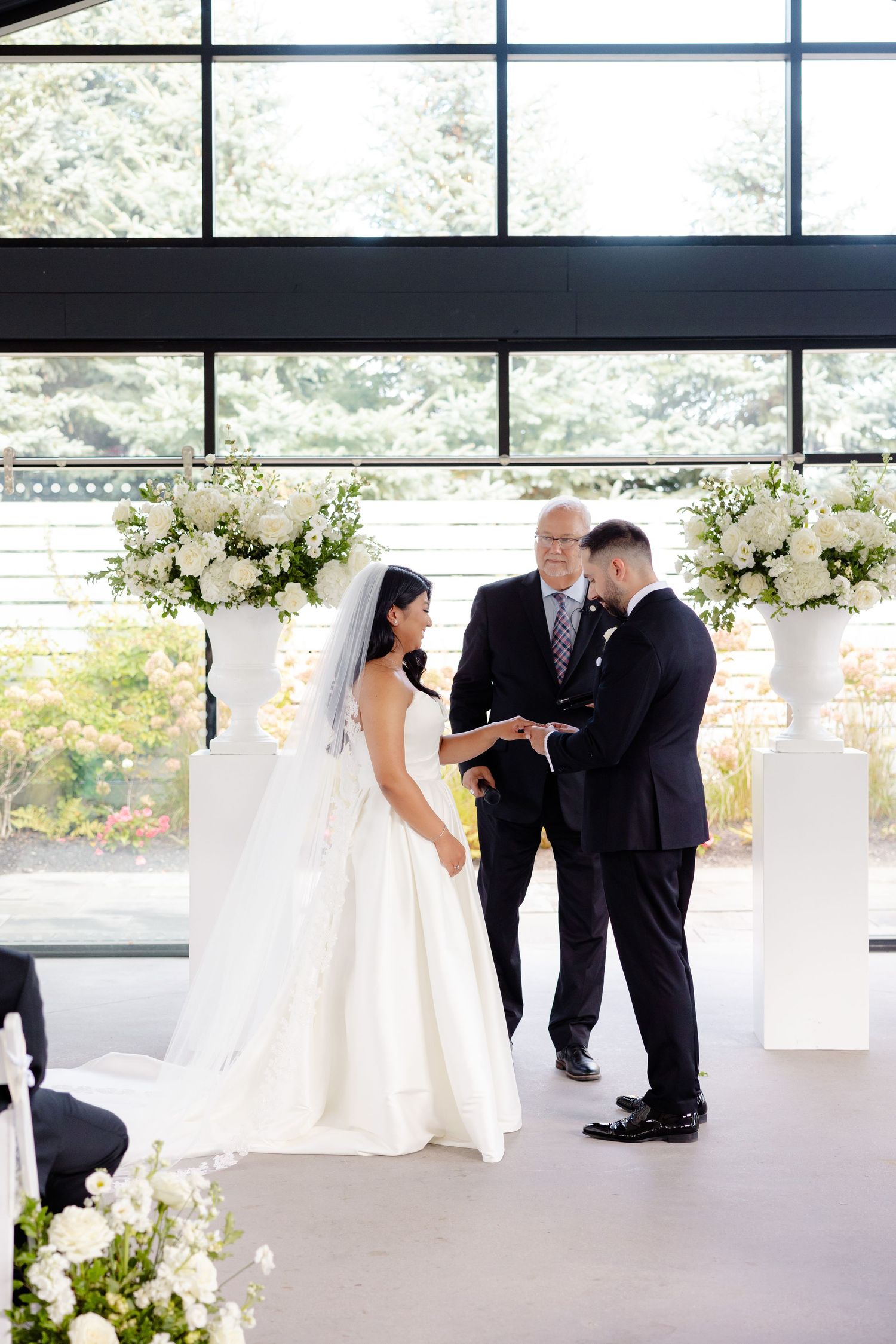 An intimate wedding ceremony moment captured in a modern venue with white floral arrangements and floor-to-ceiling windows.