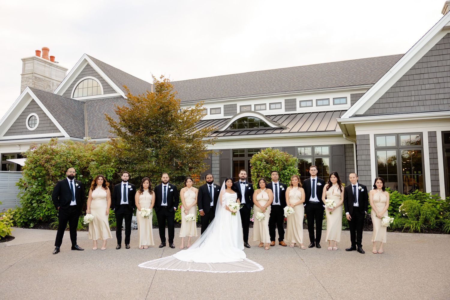 Wedding party forms V-formation pose outside gray coastal-style venue with metal roof details.