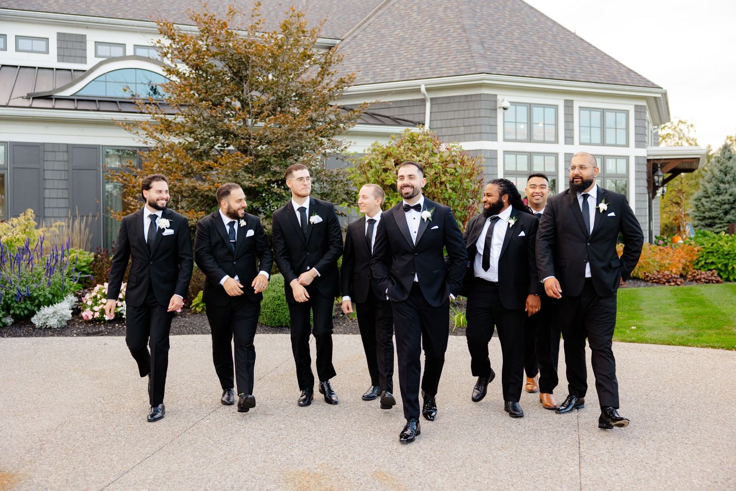 Wedding party in matching black tuxedos walking and smiling together outside elegant venue.
