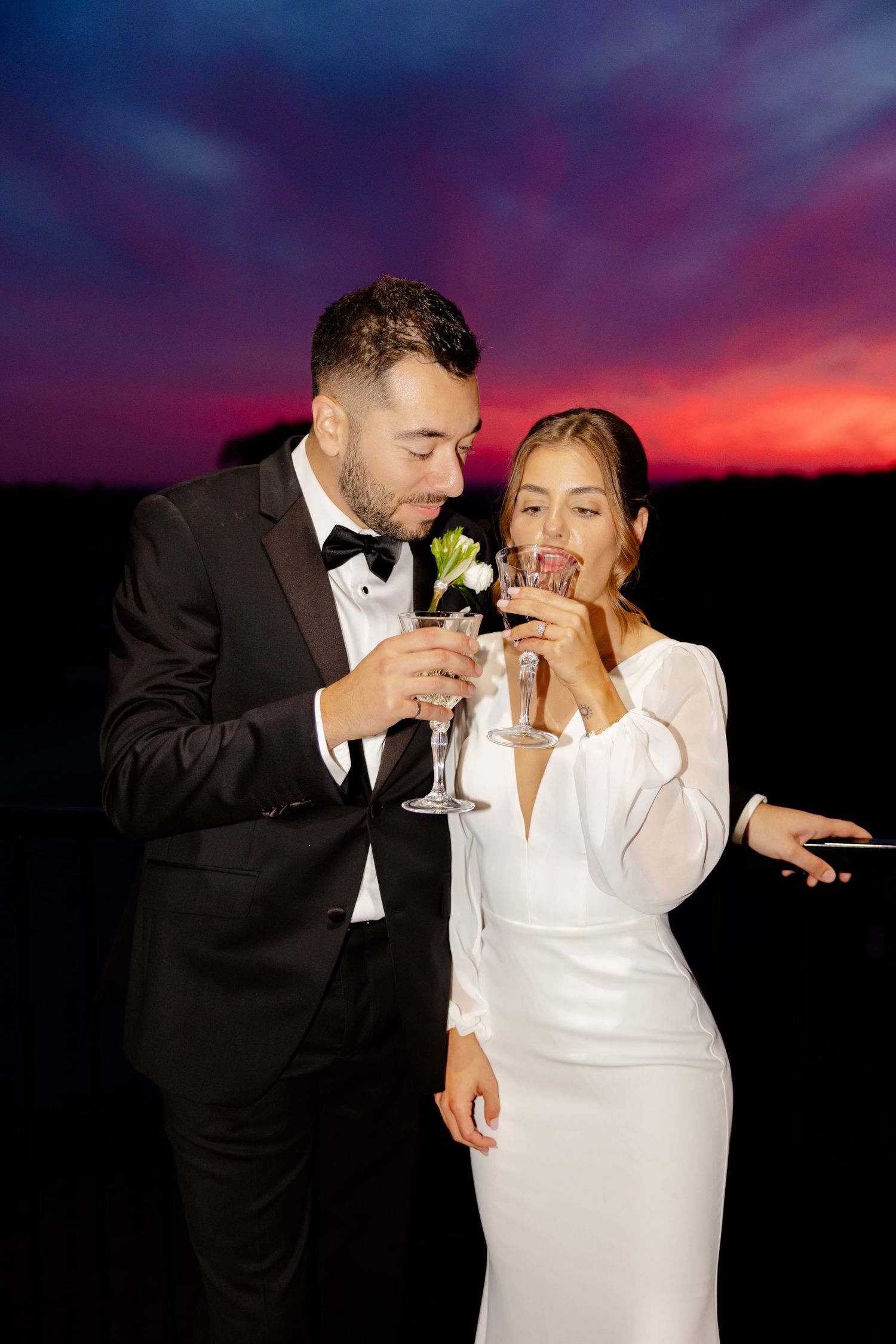 A couple in wedding attire shares a champagne toast on a deck at sunset.