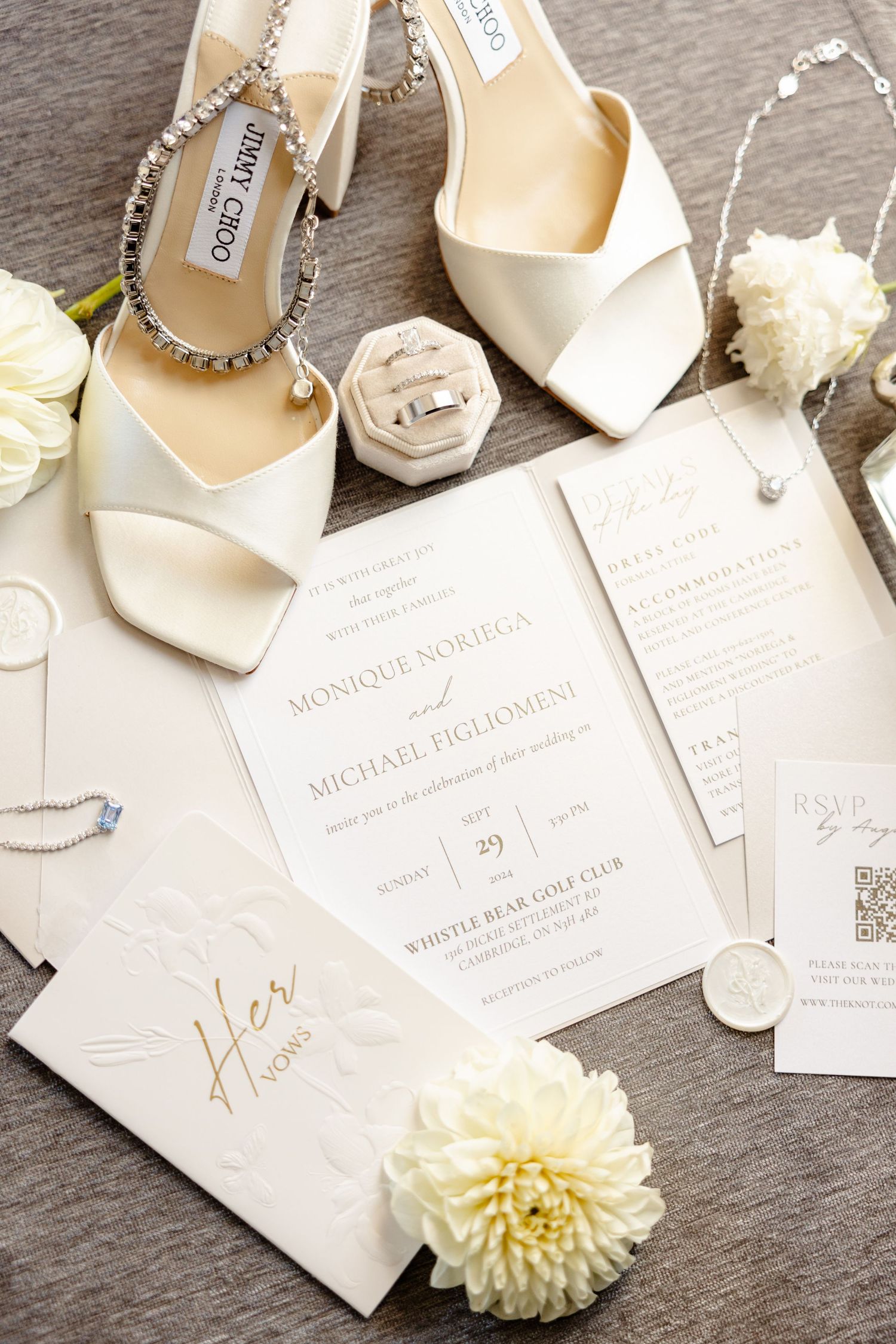White wedding invitations styled with crystal-embellished shoes and cream florals on gray background.
