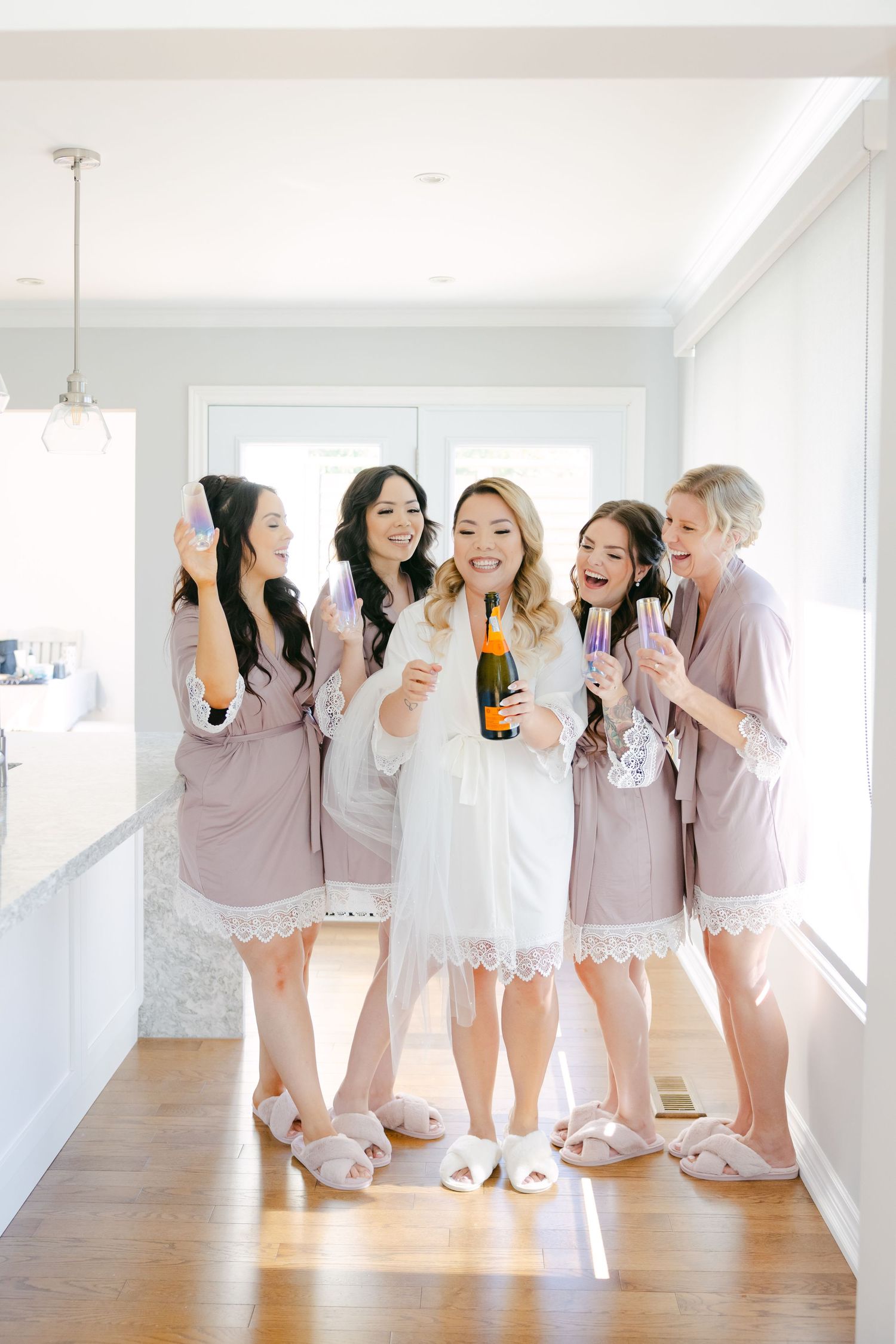 Bridesmaids in blush robes celebrating with champagne in bright modern kitchen.