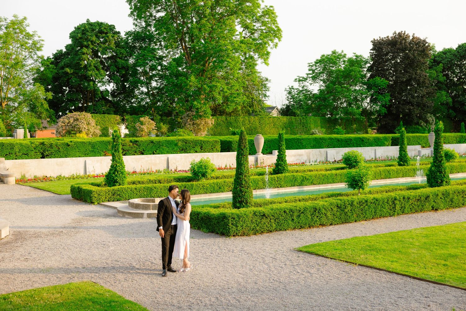 A couple walks through a formal garden with perfectly trimmed hedges and cypress trees.