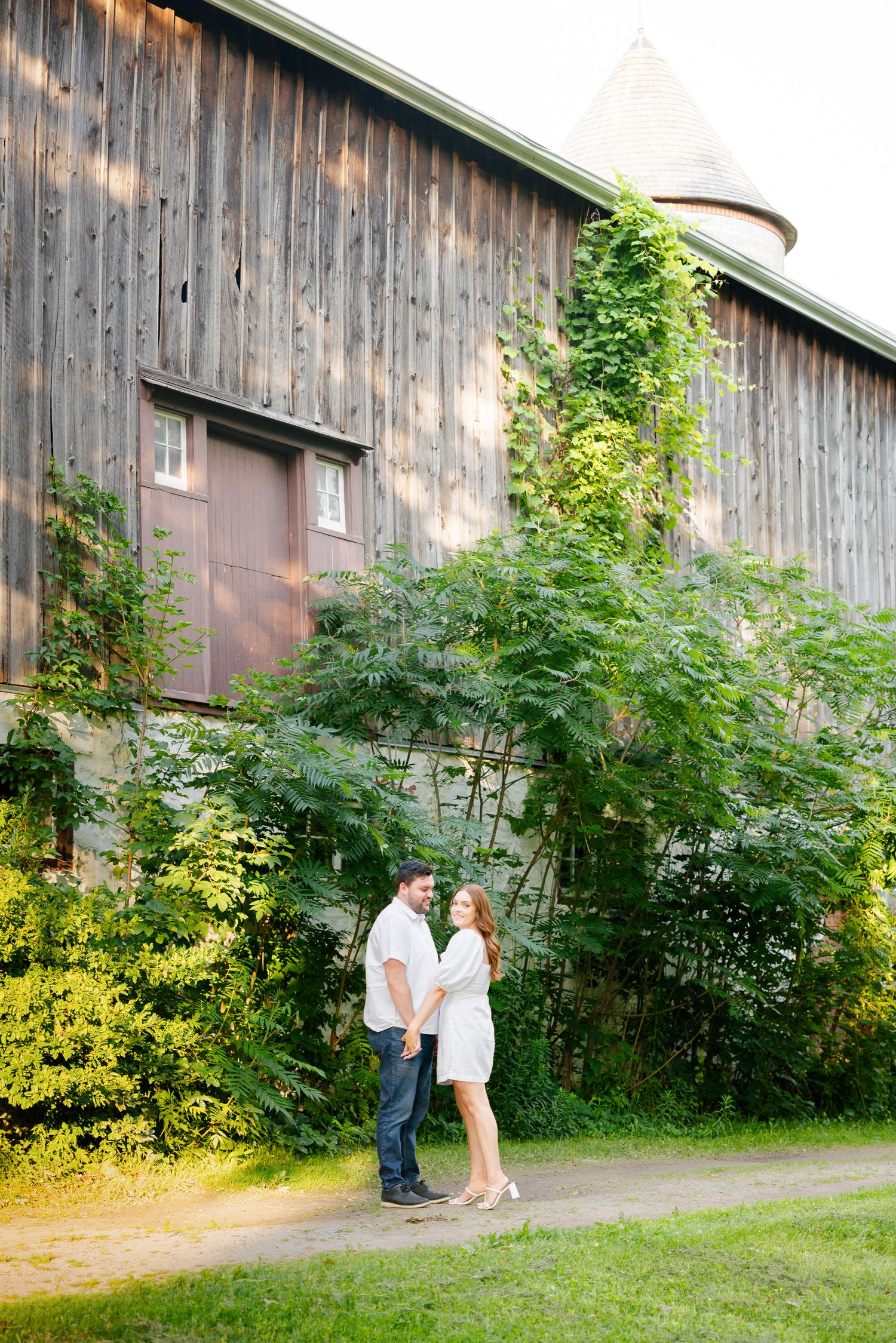 A tender moment between a couple standing near a rustic barn with lush summer foliage.