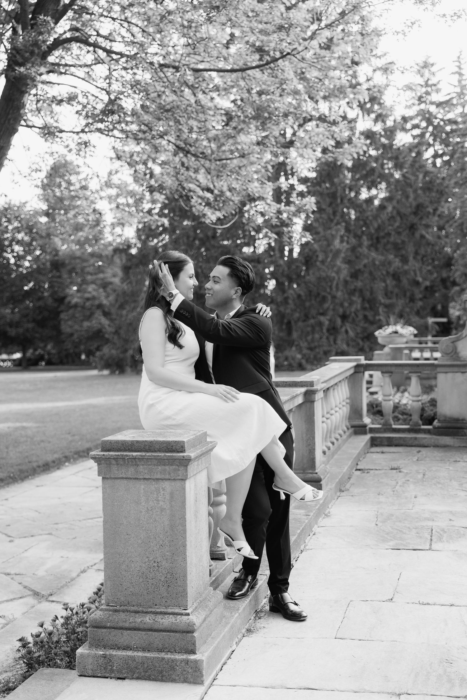 Black and white photo of couple posed romantically on stone railing in garden setting.