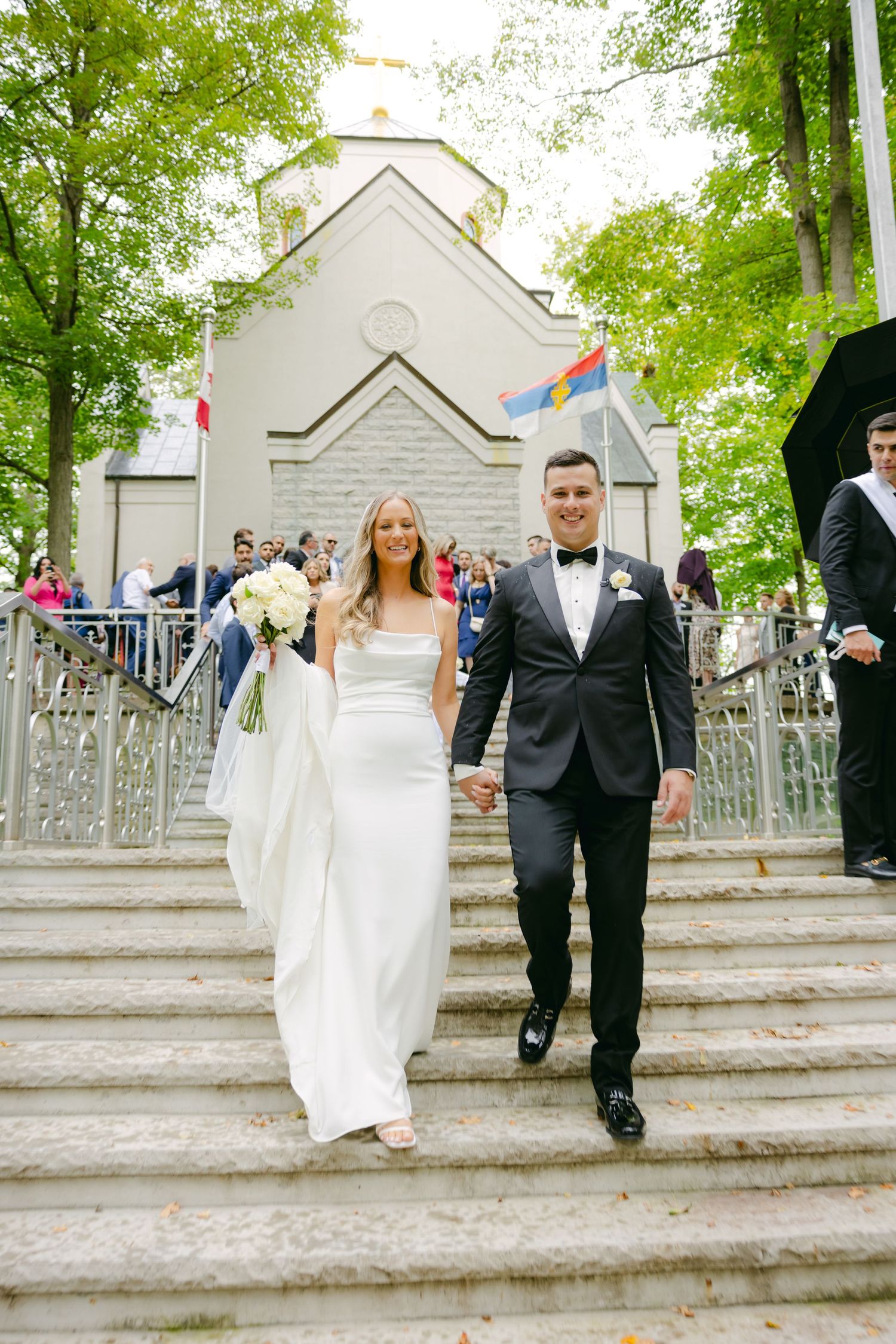 Newlyweds descend white stone steps outside a charming white church with trees in background.