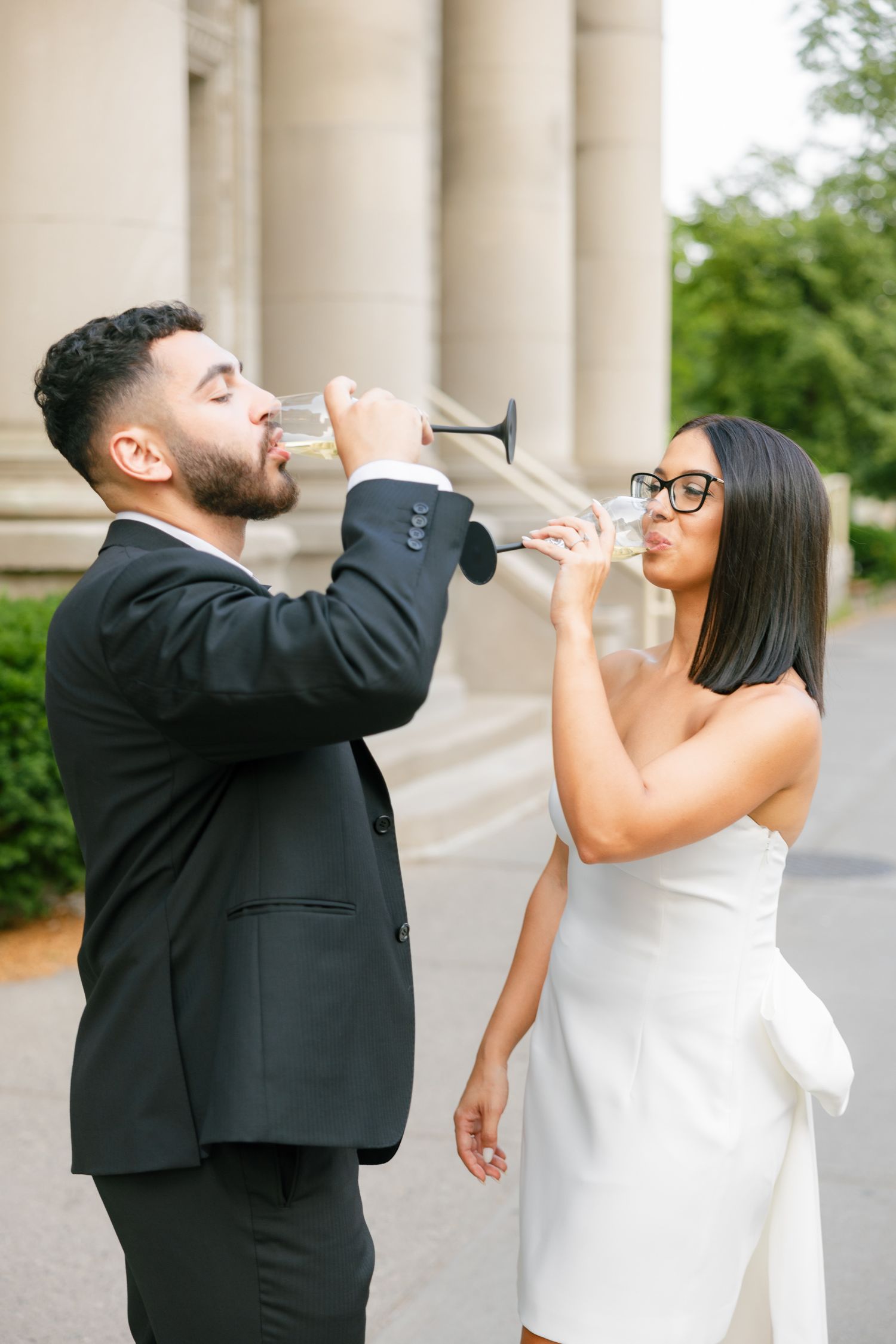 Playful moment as couple in formal attire drink champagne together by stone pillars.