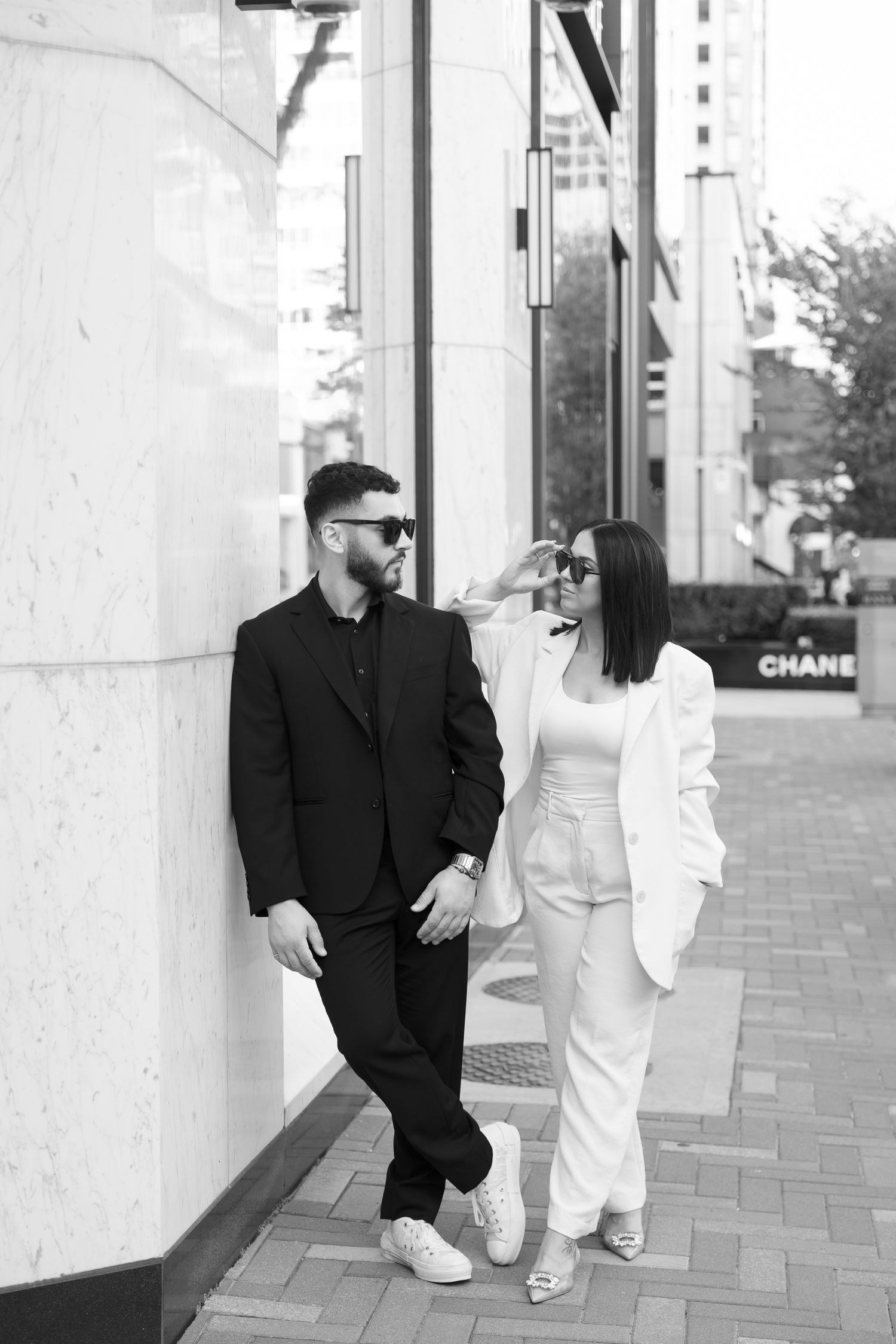 Black and white photo of couple in matching formal attire posing against white building wall.