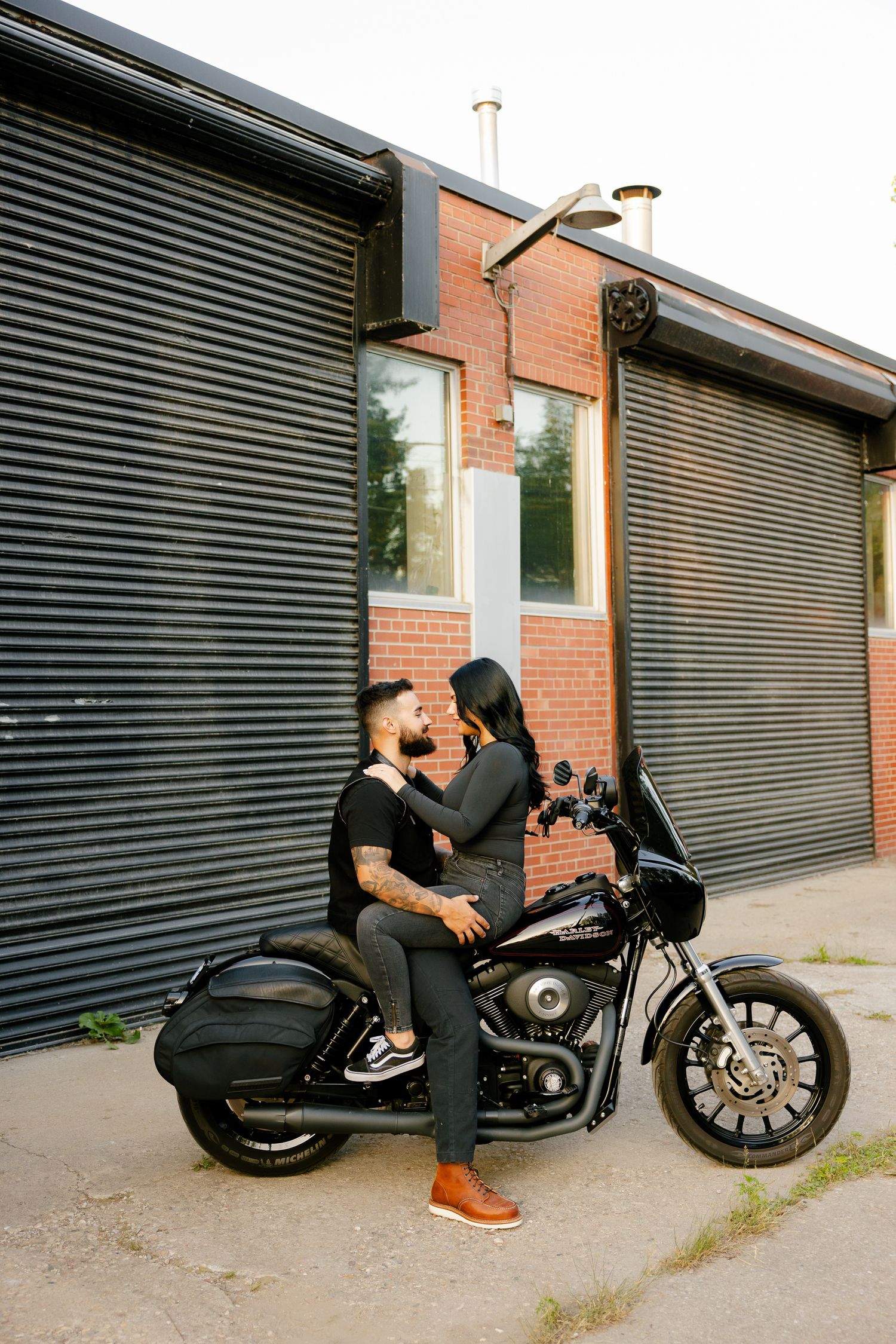 A couple embraces while seated on their parked motorcycle near a brick building.