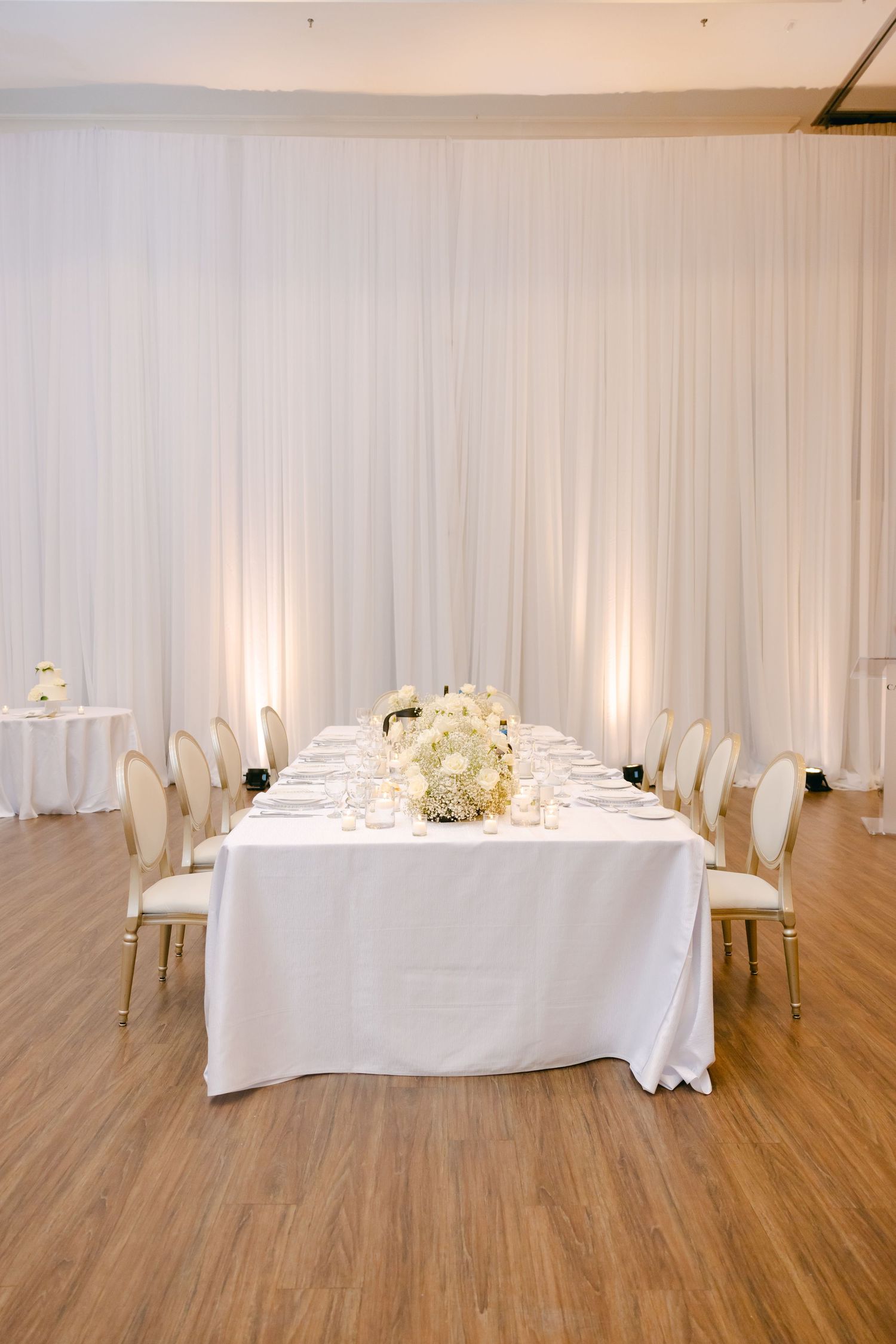 Minimalist wedding reception setup with white draping, long table, and cream chairs in a bright venue.