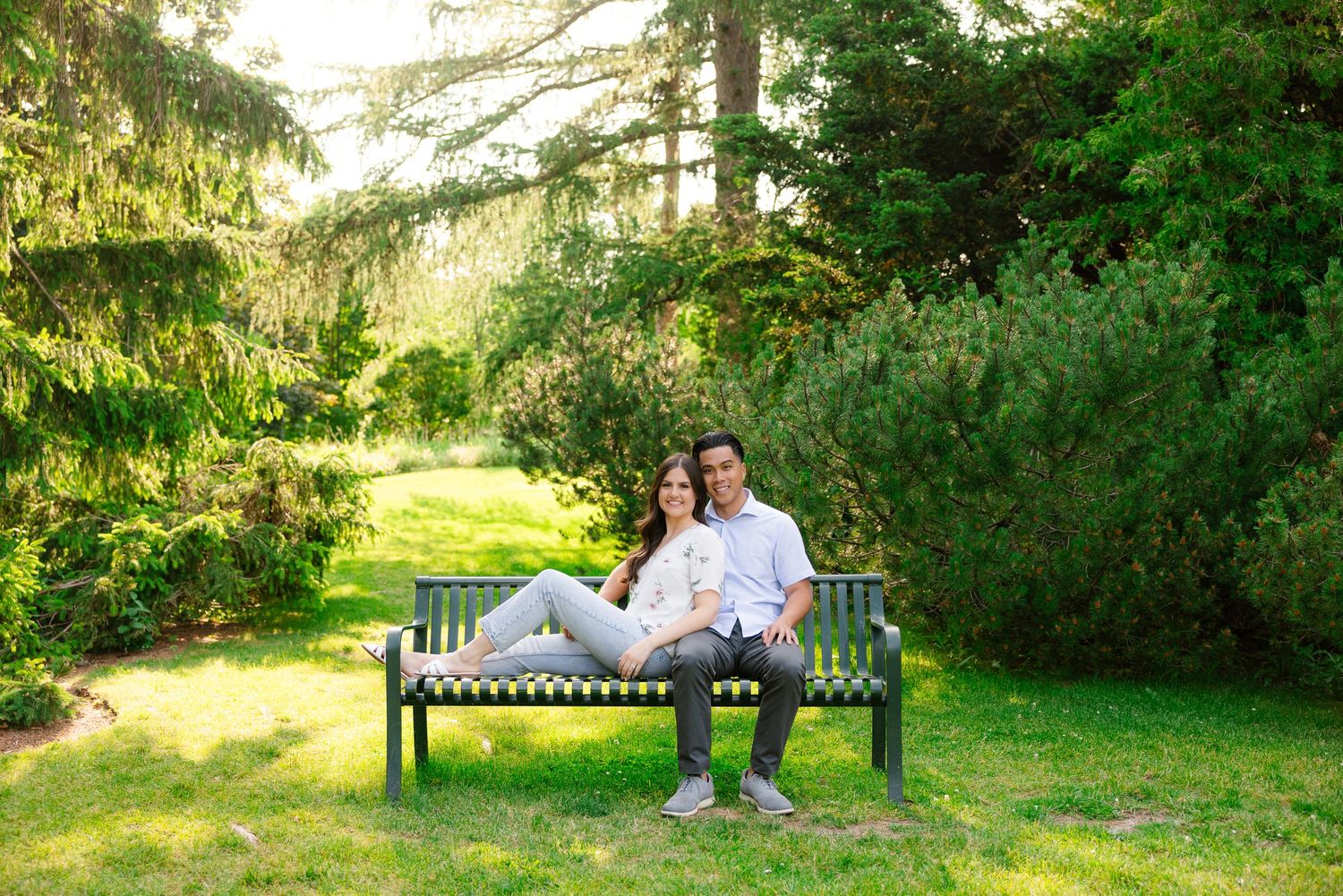 A couple relaxes together on a garden bench surrounded by evergreen trees and summer foliage.