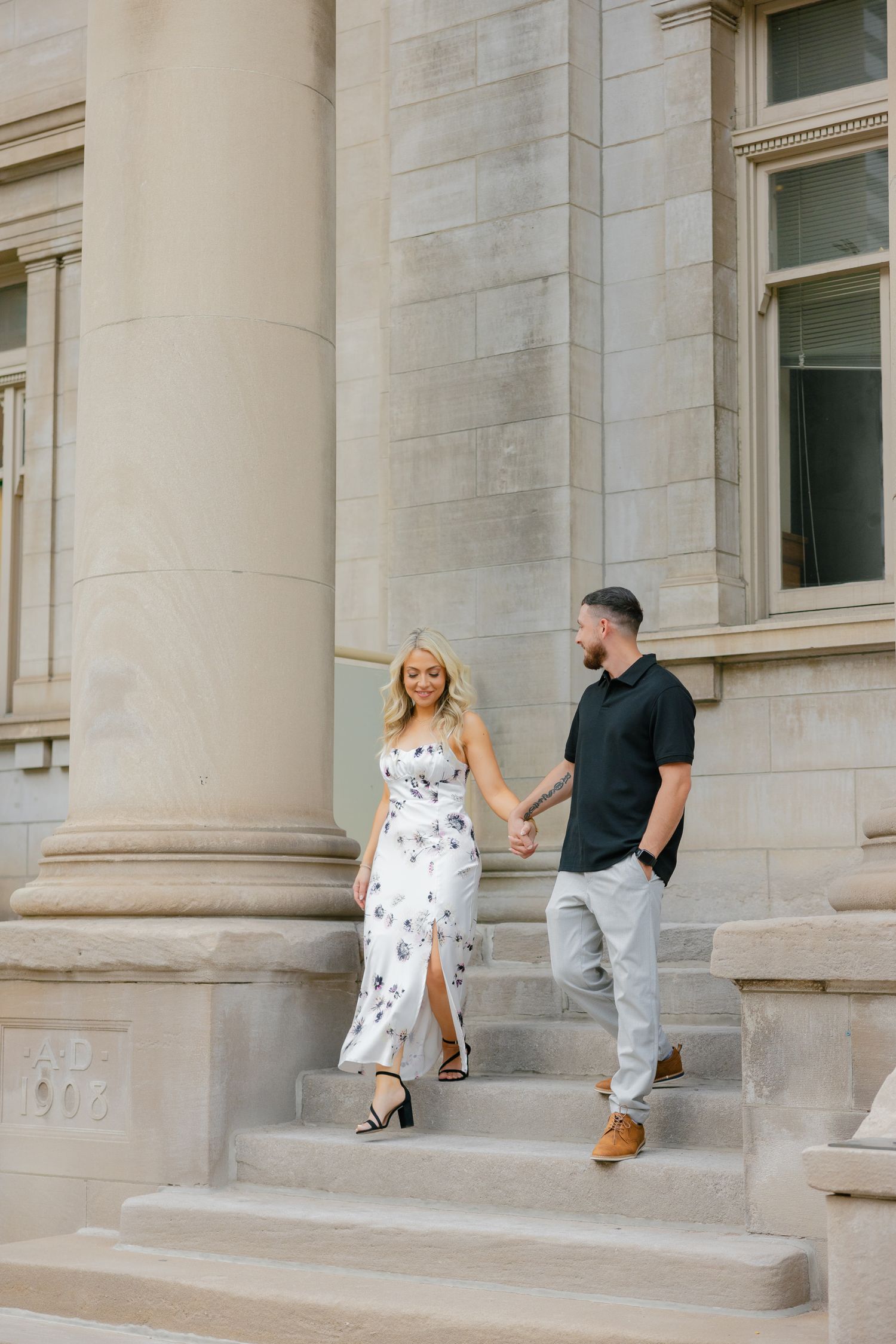 A couple descends elegant stone steps while holding hands beside a classical building column.