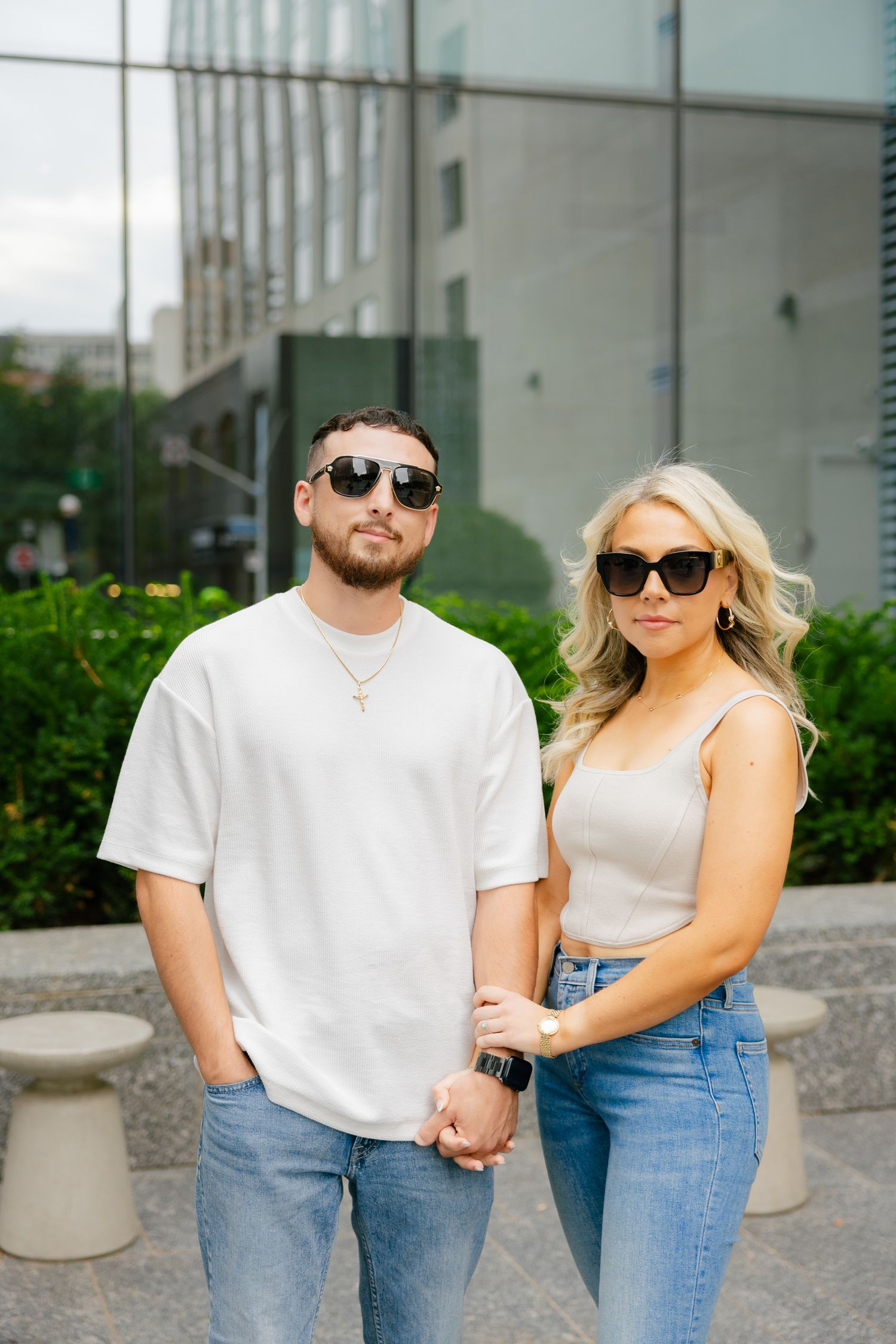 A couple in matching denim and sunglasses stand together near urban landscaping.