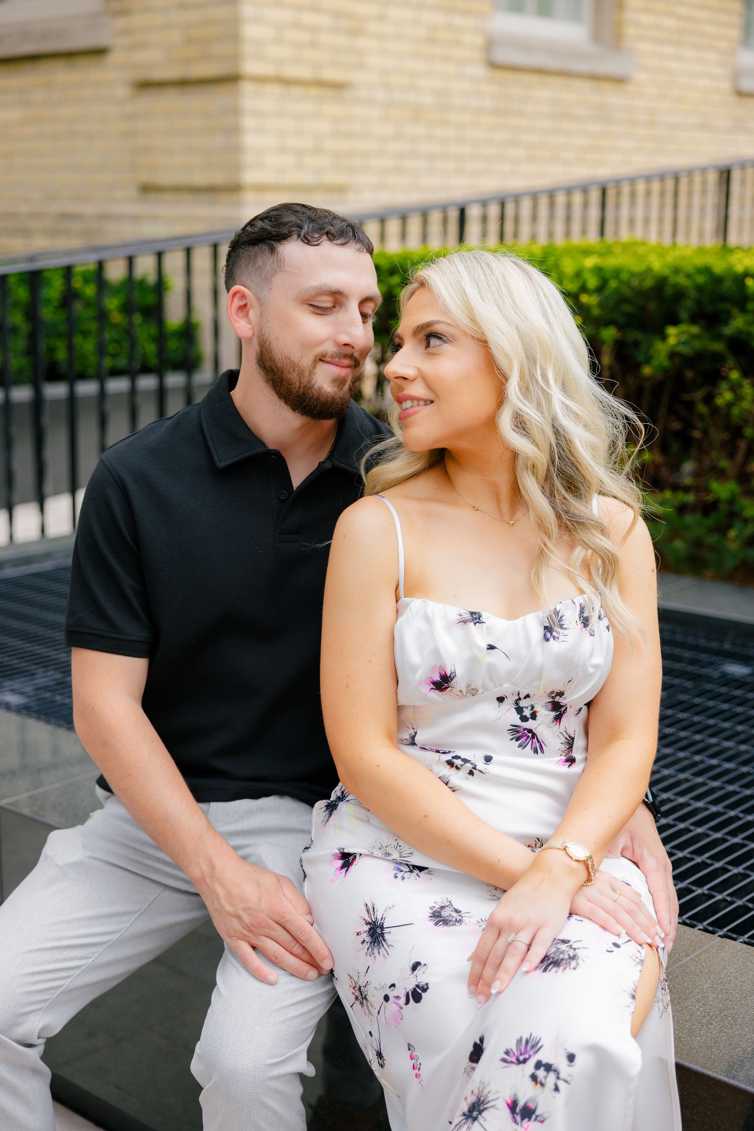 A couple sits intimately together outdoors, with her wearing a floral summer dress and him in a black polo.