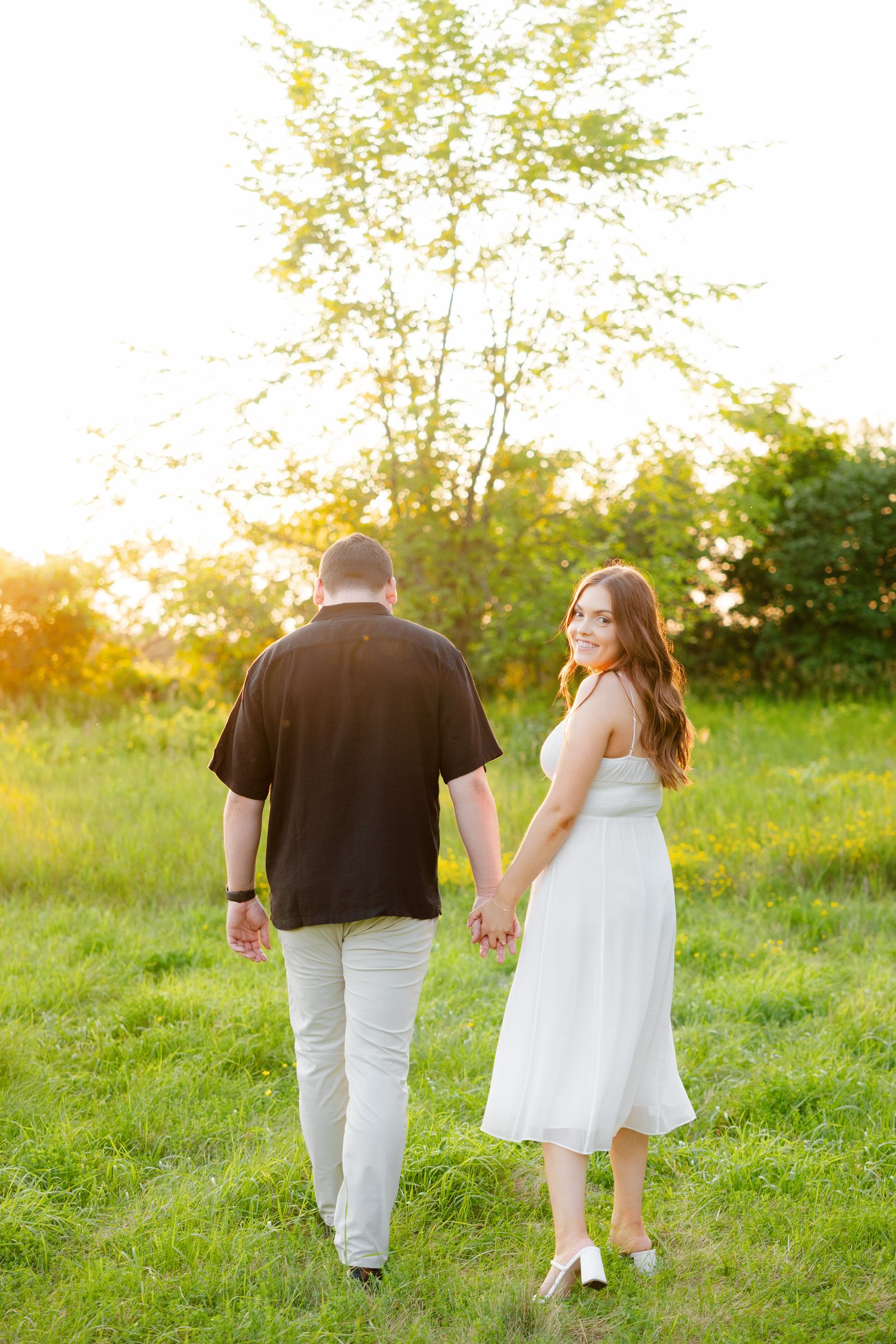 Couple walks together through sunlit meadow holding hands in coordinated outfits.