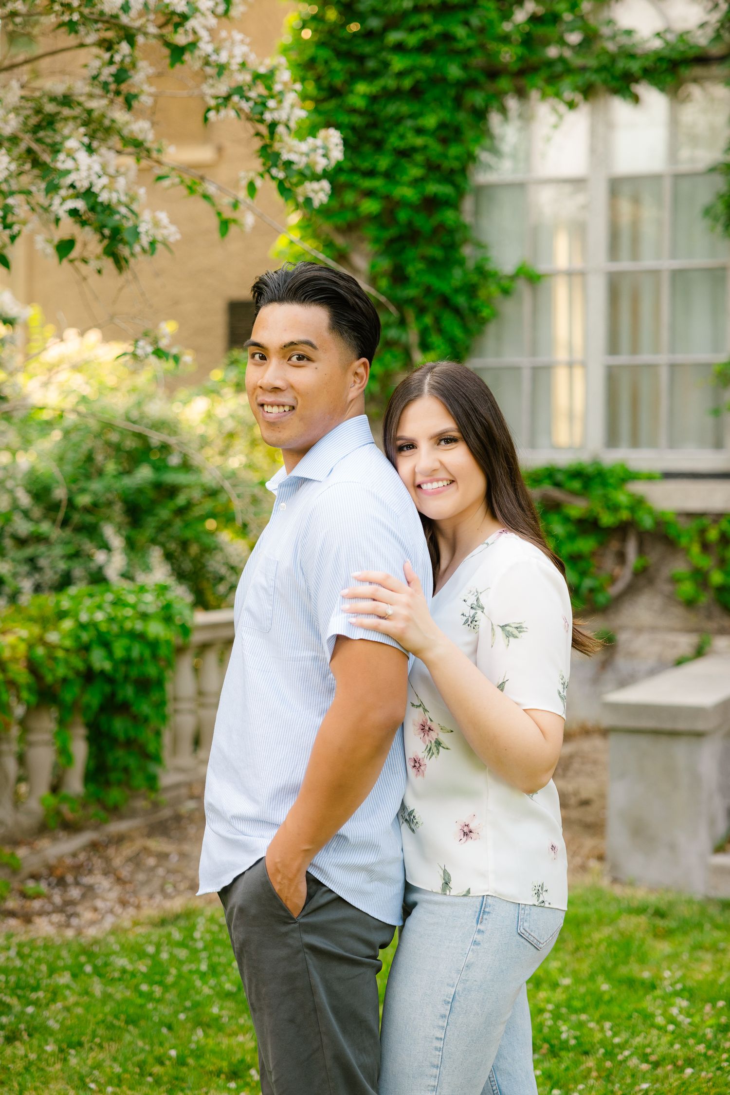 Couple smiles while posing against green ivy and flowering tree backdrop.