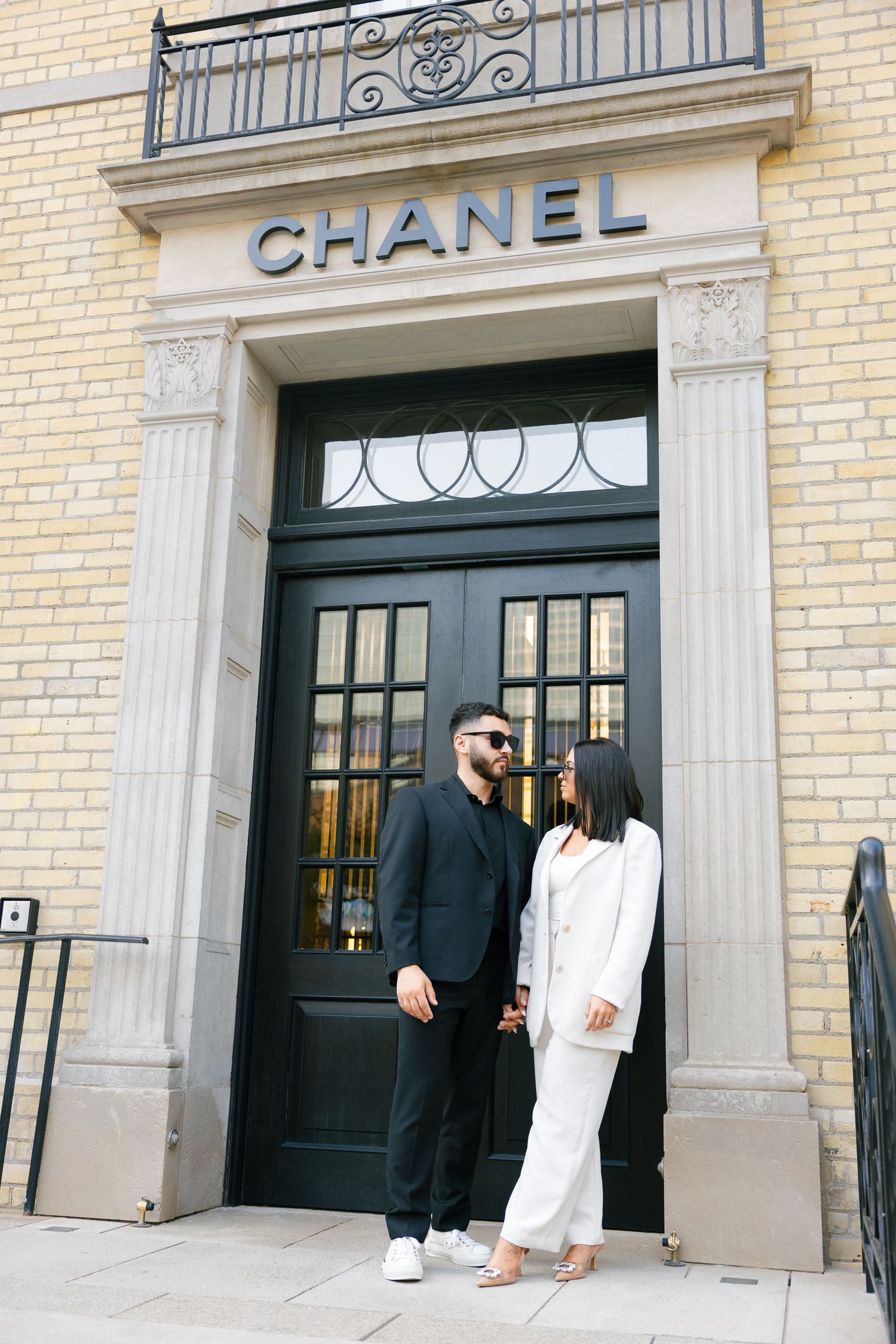 A stylish duo poses against the iconic Chanel storefront with ornate black doors.