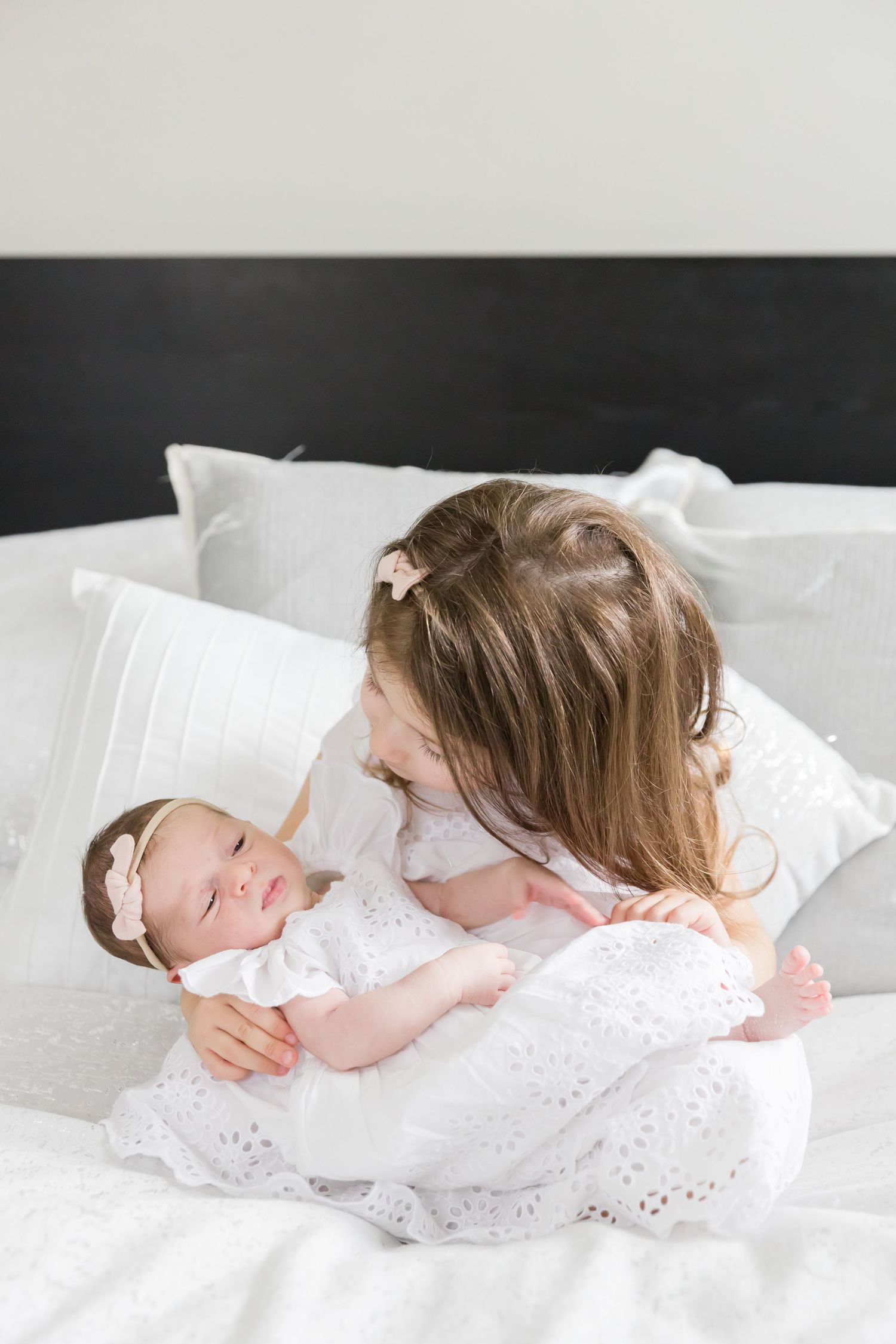 Two children in white dresses sharing tender moment on white bedding.