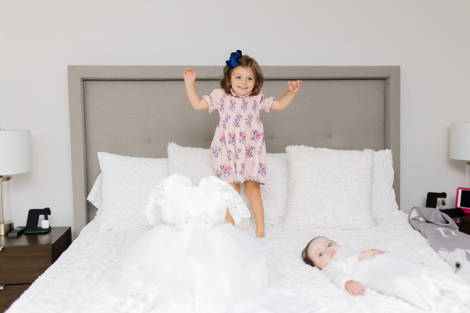 A child in pink jumps on a white bed while another rests nearby.