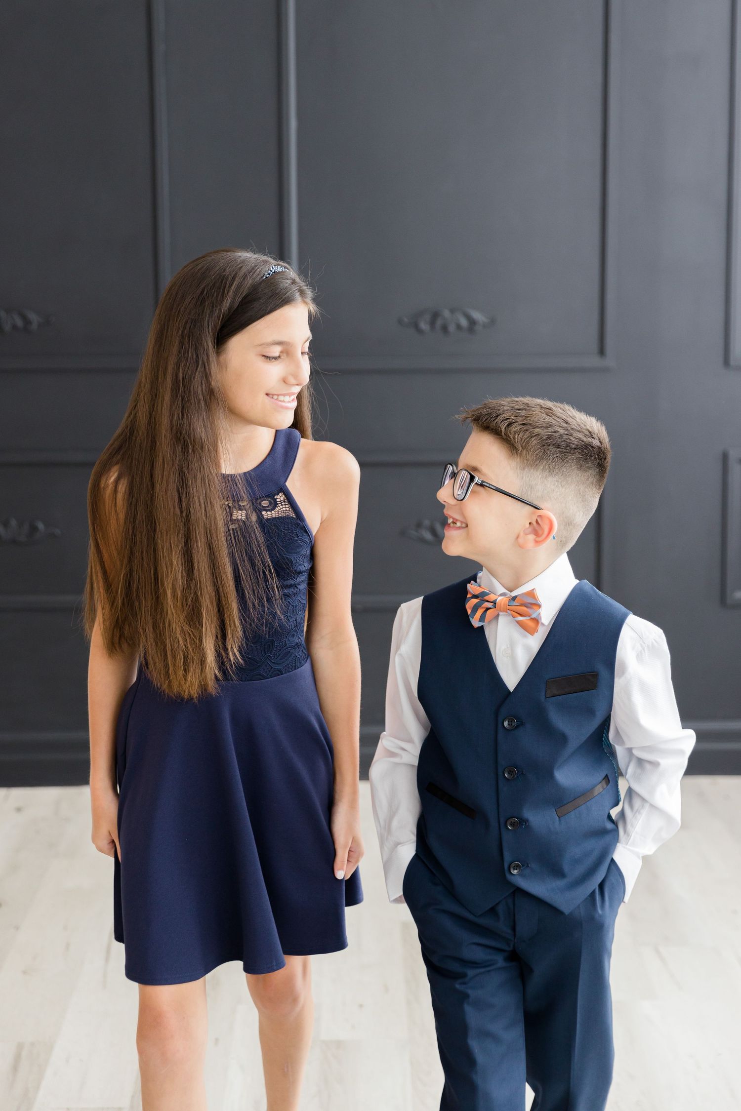 Siblings in coordinating navy formal wear share a sweet moment against a gray paneled wall.