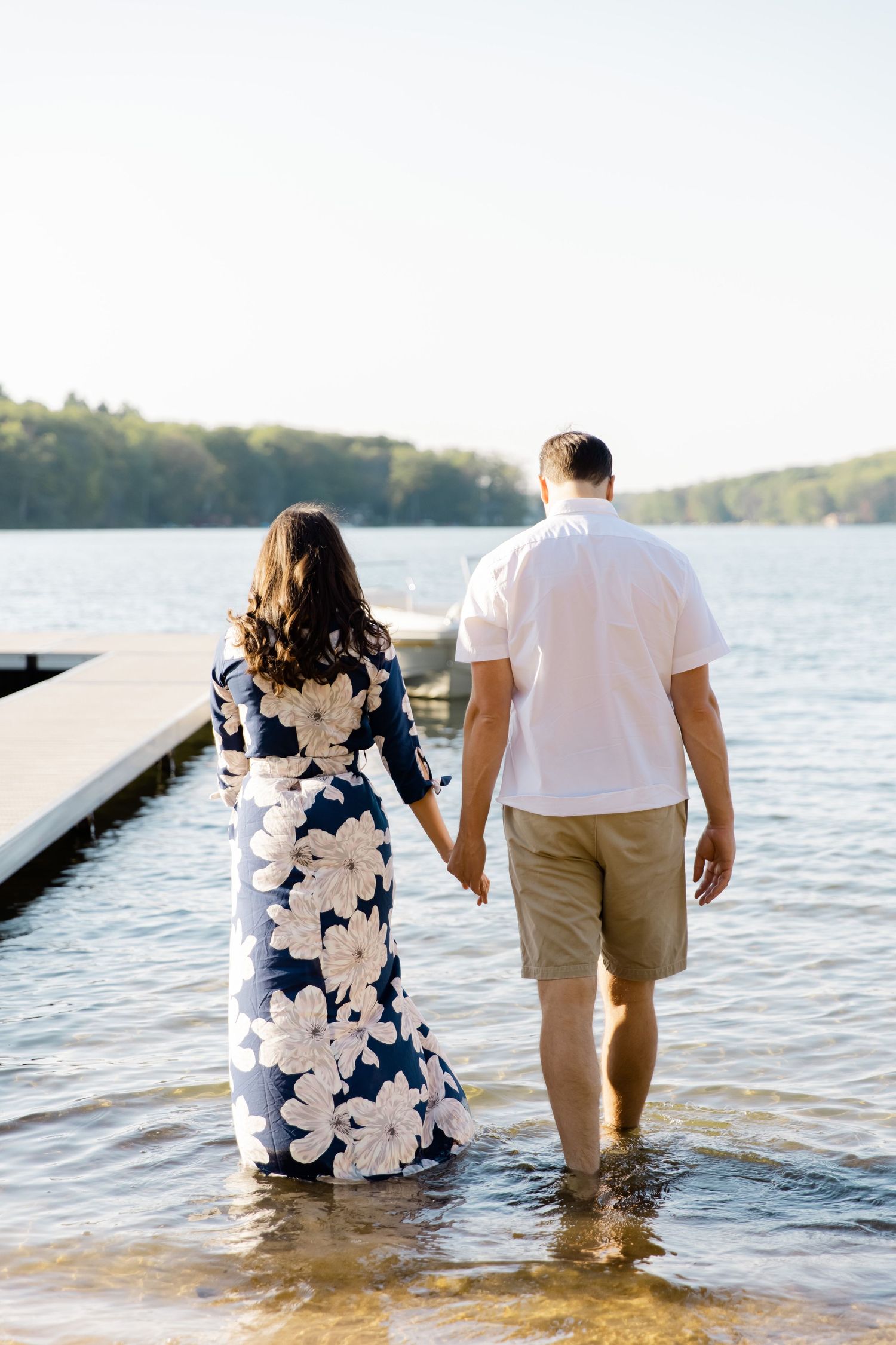 A couple holding hands walks through shallow water near a dock with a floral dress and casual summer attire.