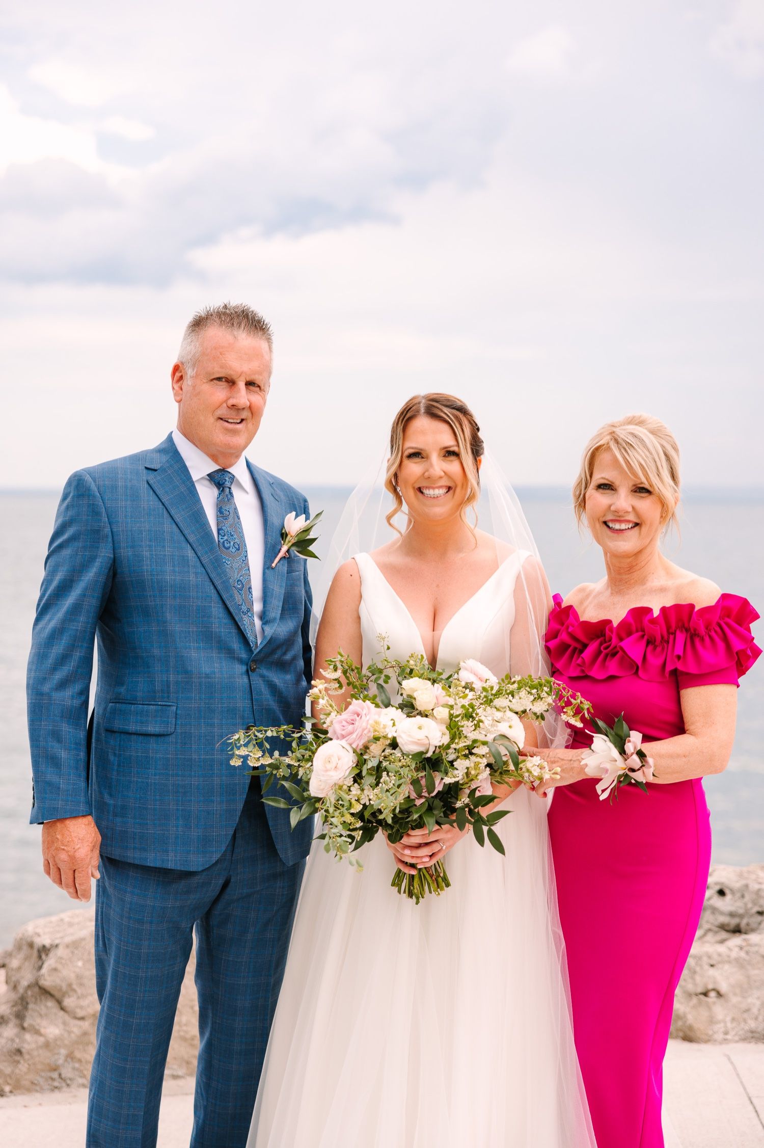 Wedding party members pose together against a lakefront backdrop on a cloudy day.