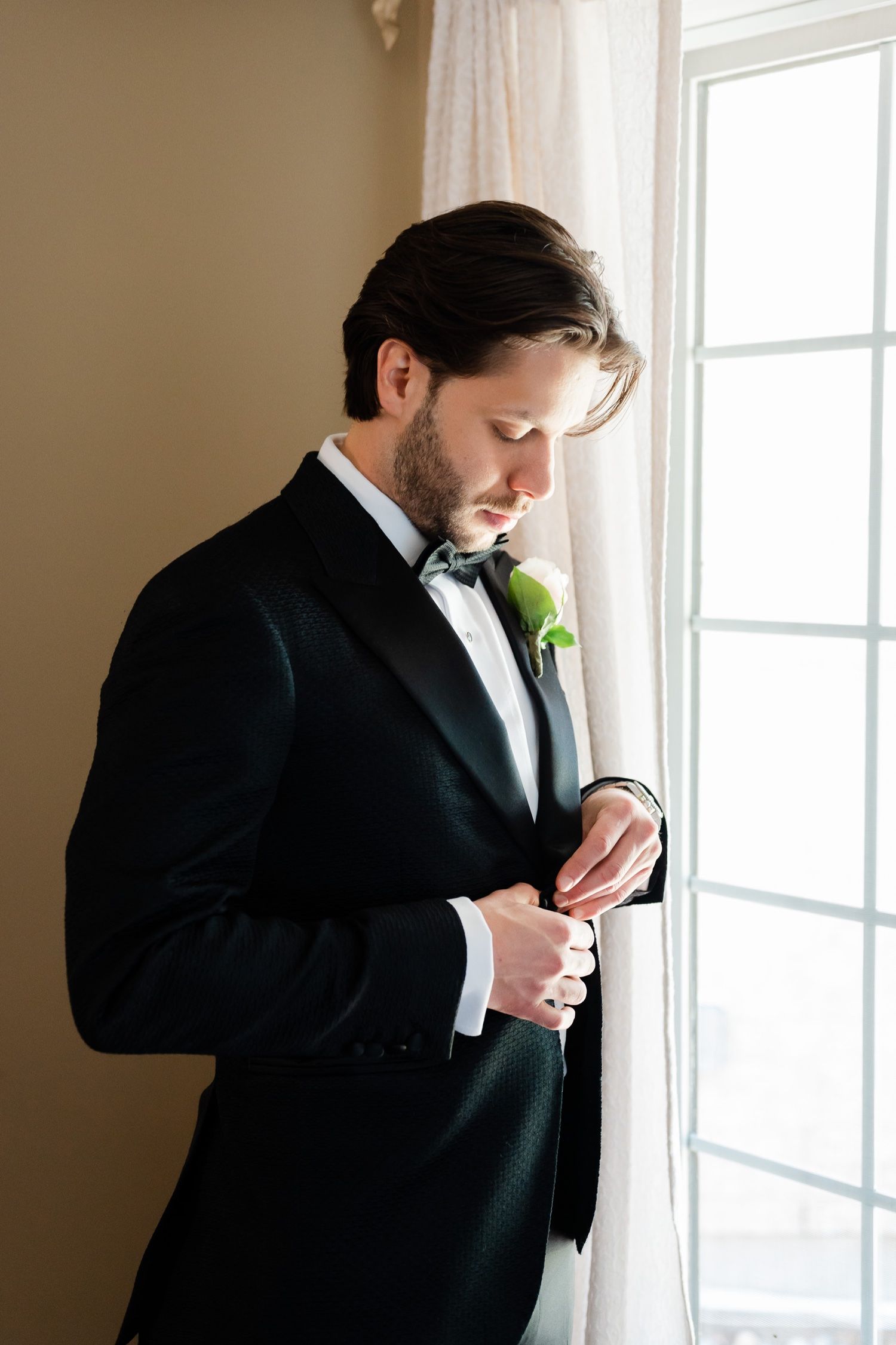 Close-up shot of a person in black tuxedo and boutonniere adjusting cuffs by window.