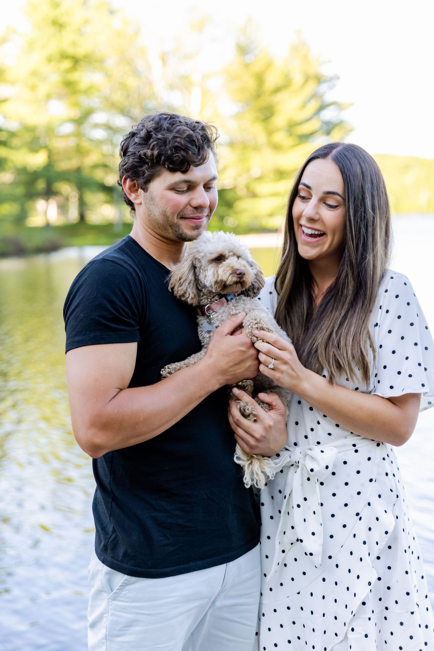Couple holding small fluffy dog while posing by lake in summer attire.