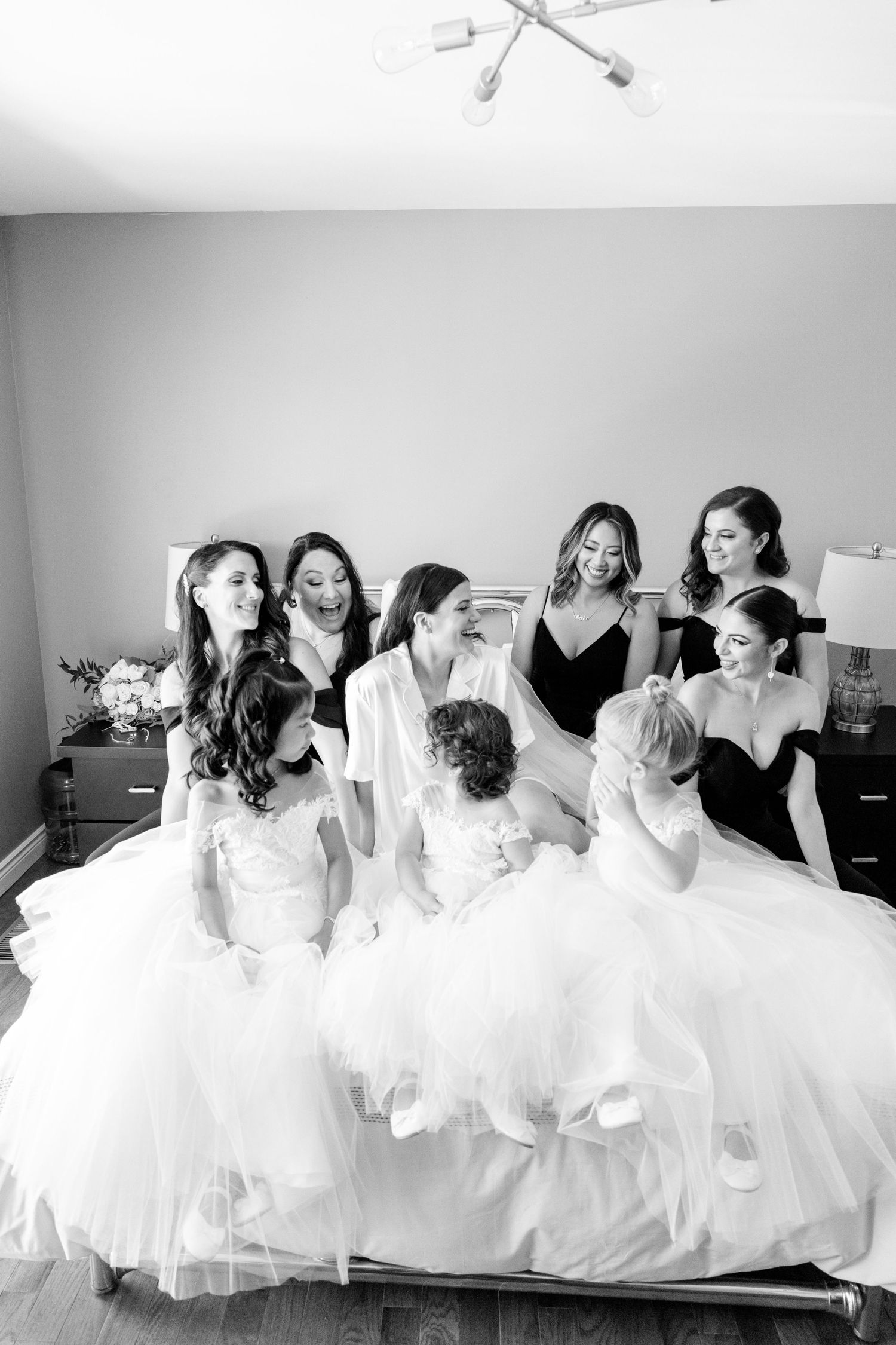 Black and white candid moment of bridal party gathered on bed sharing laughter.