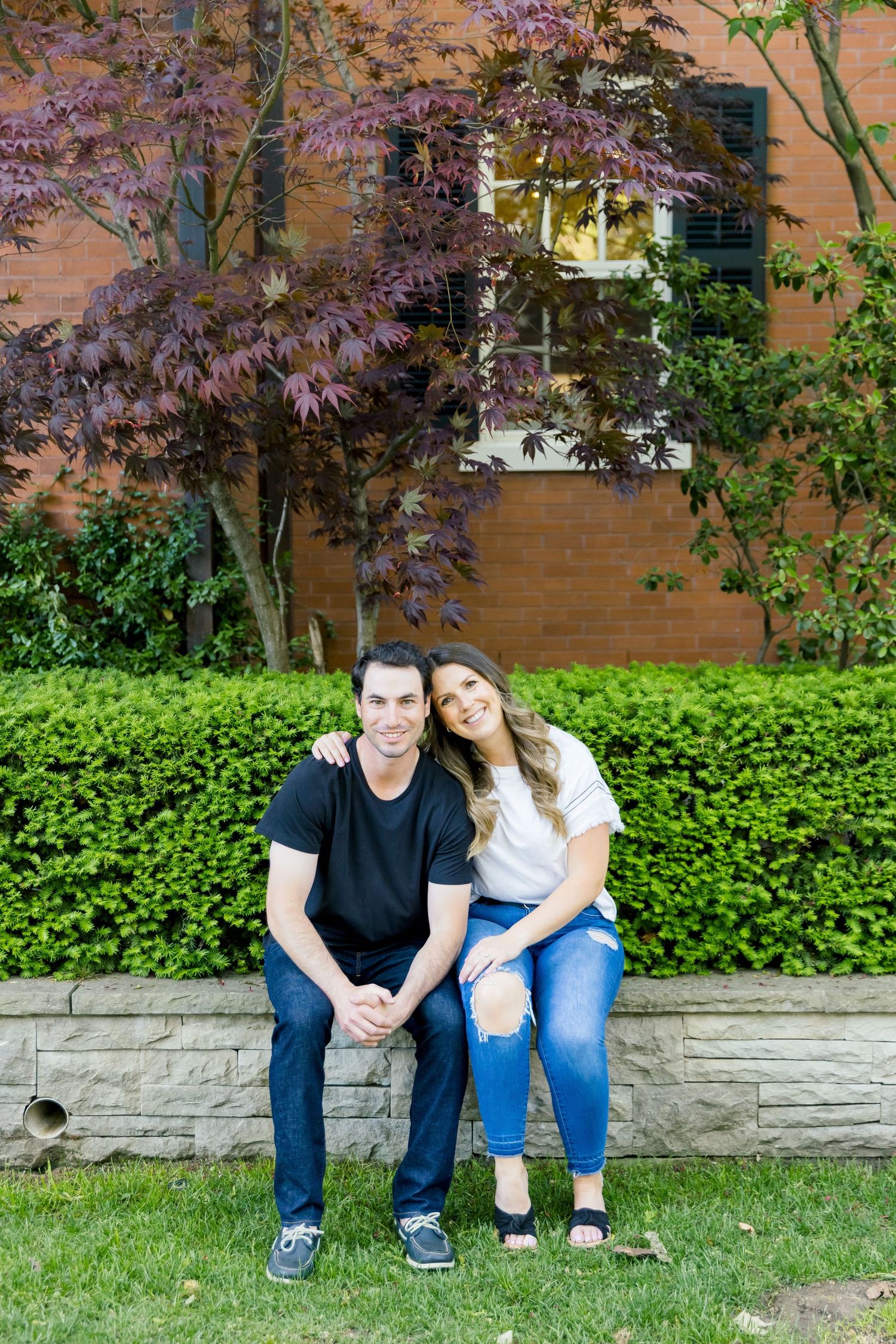 A couple relaxes on a stone retaining wall with Japanese maple and brick building backdrop.
