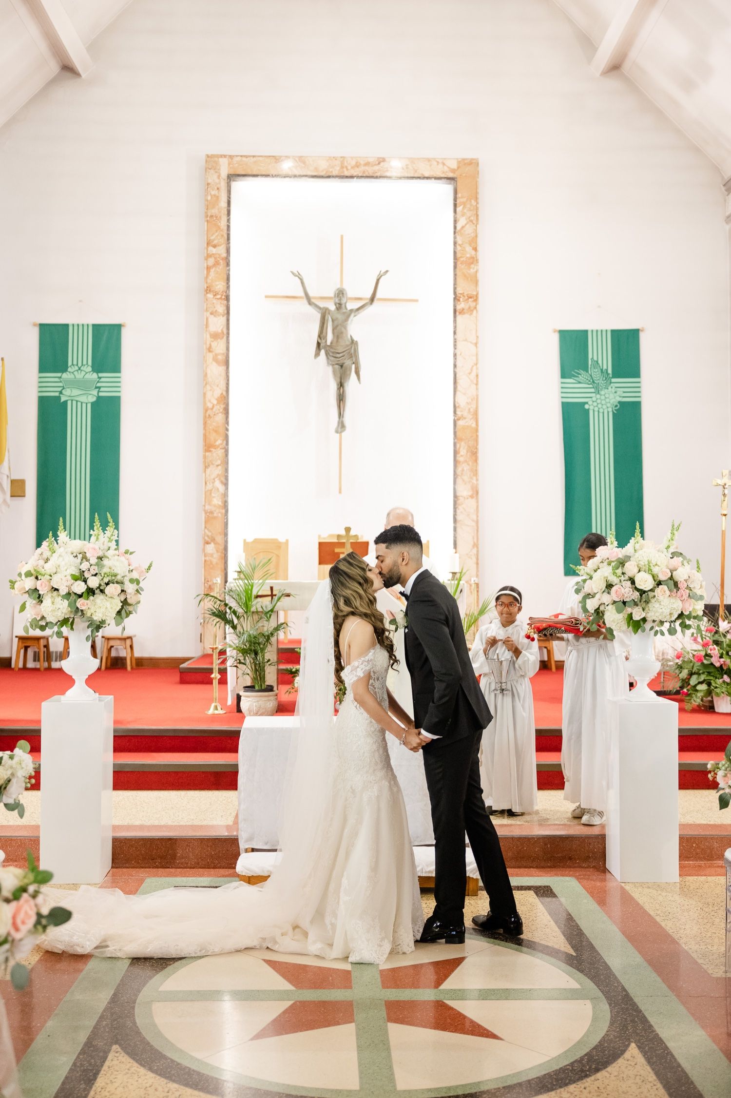 A wedding ceremony in a white church with green banners and floral arrangements lining the aisle.