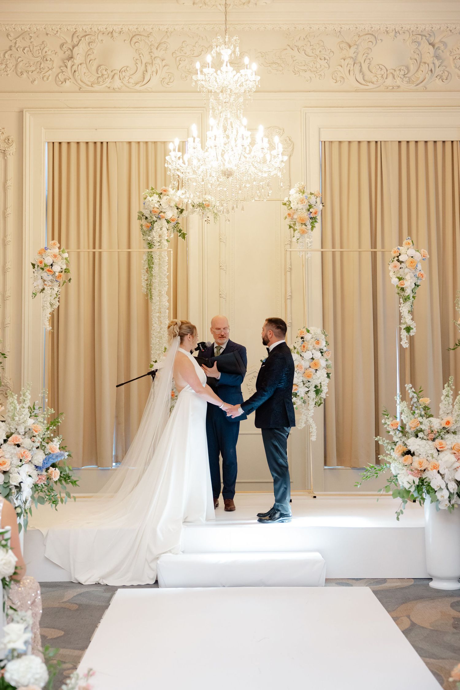 Elegant wedding ceremony setup with crystal chandelier and floral arch in a cream-colored ballroom.