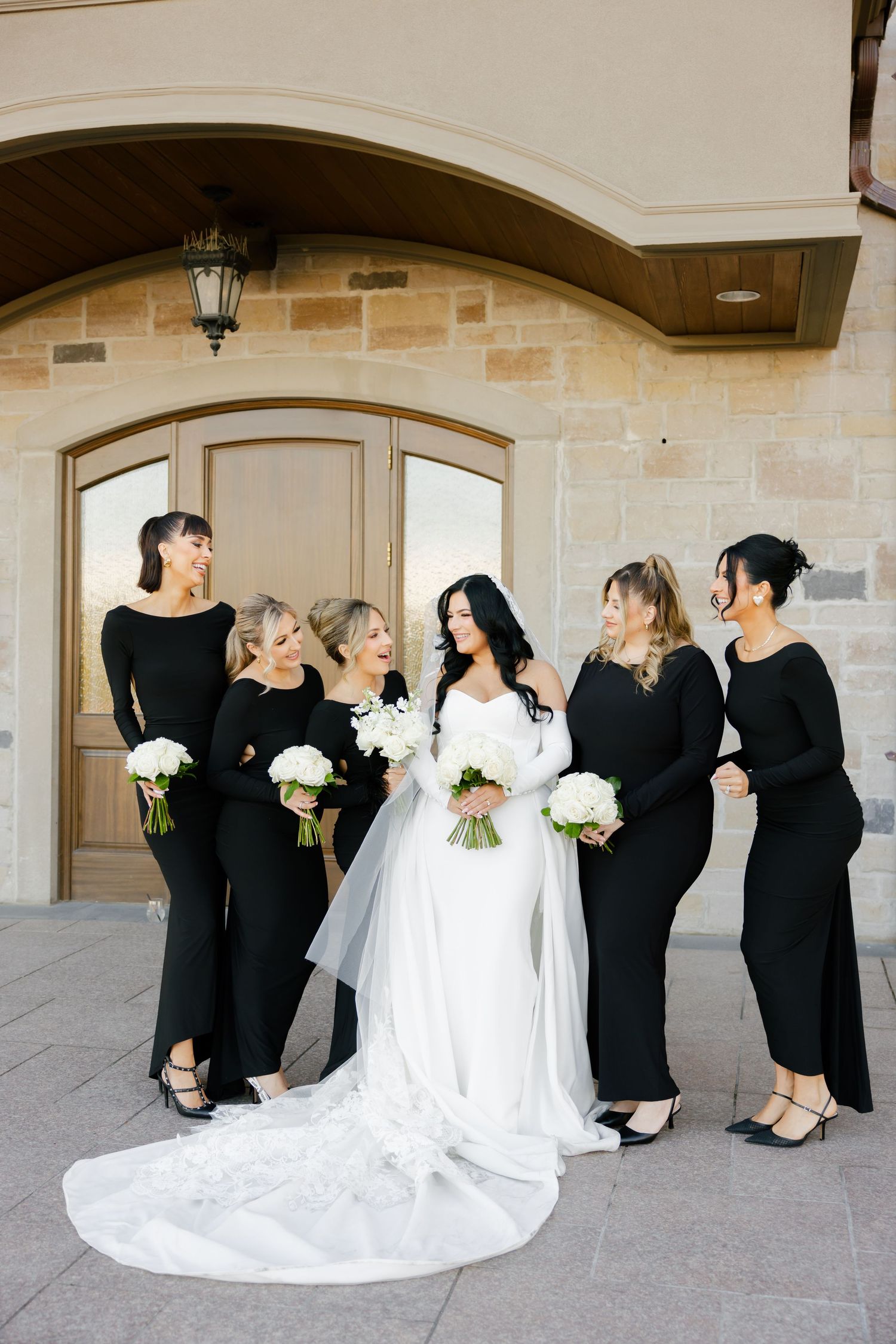 Bridal party laughs together during formal photos outside stone building.