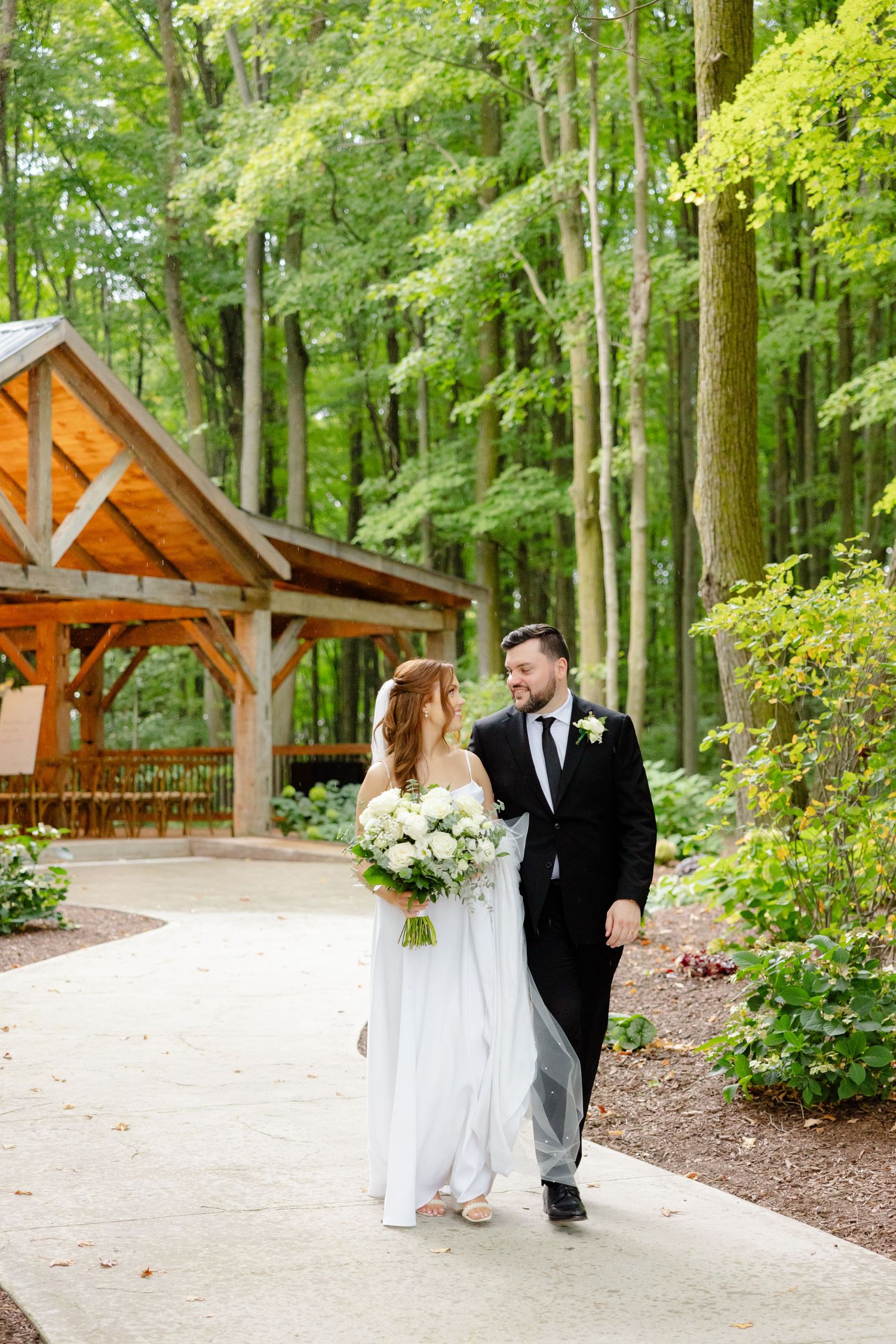 Couple walks along garden path with rustic pavilion and green forest scenery.