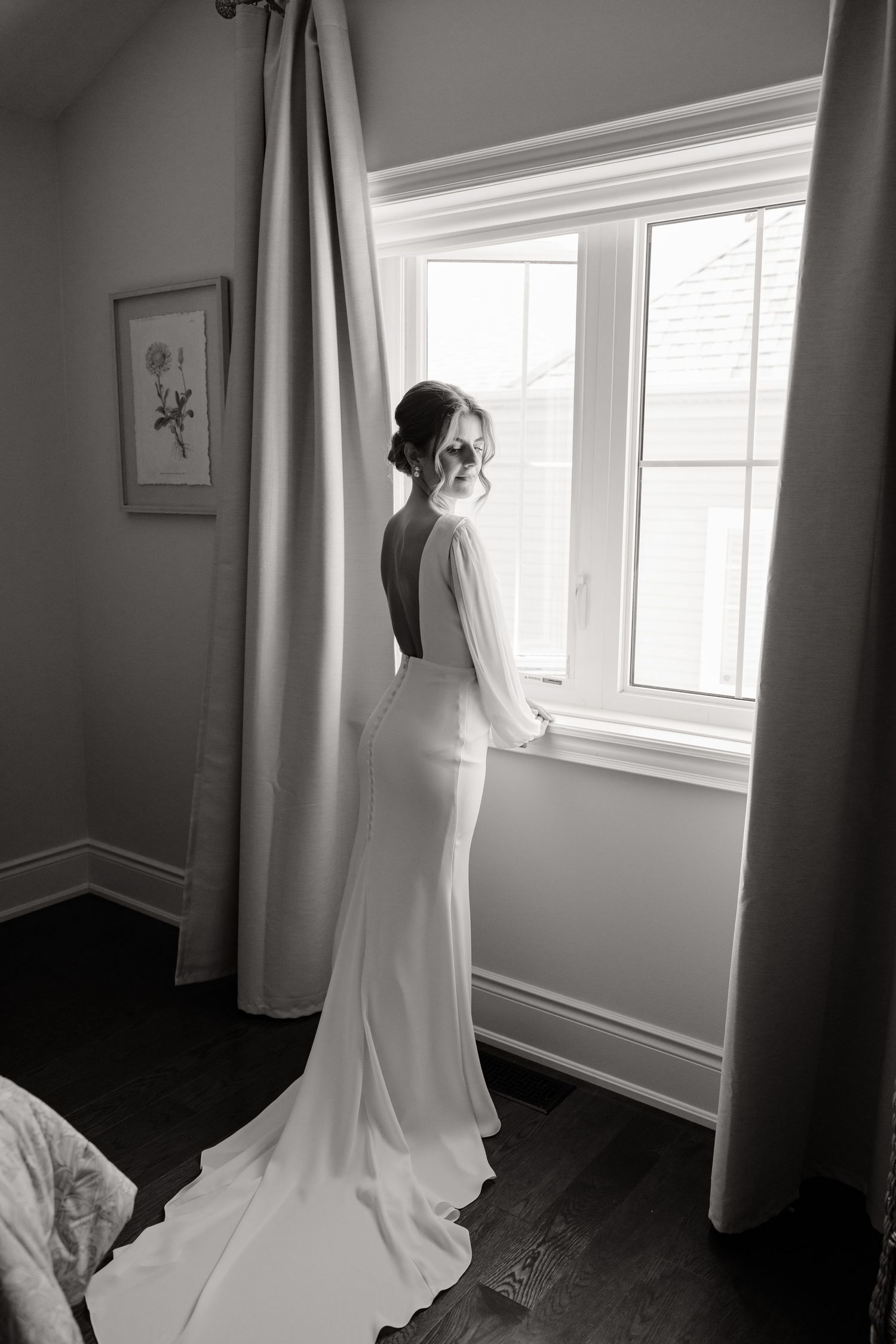 Black and white photograph of an elegant backless wedding gown with long train by a window.