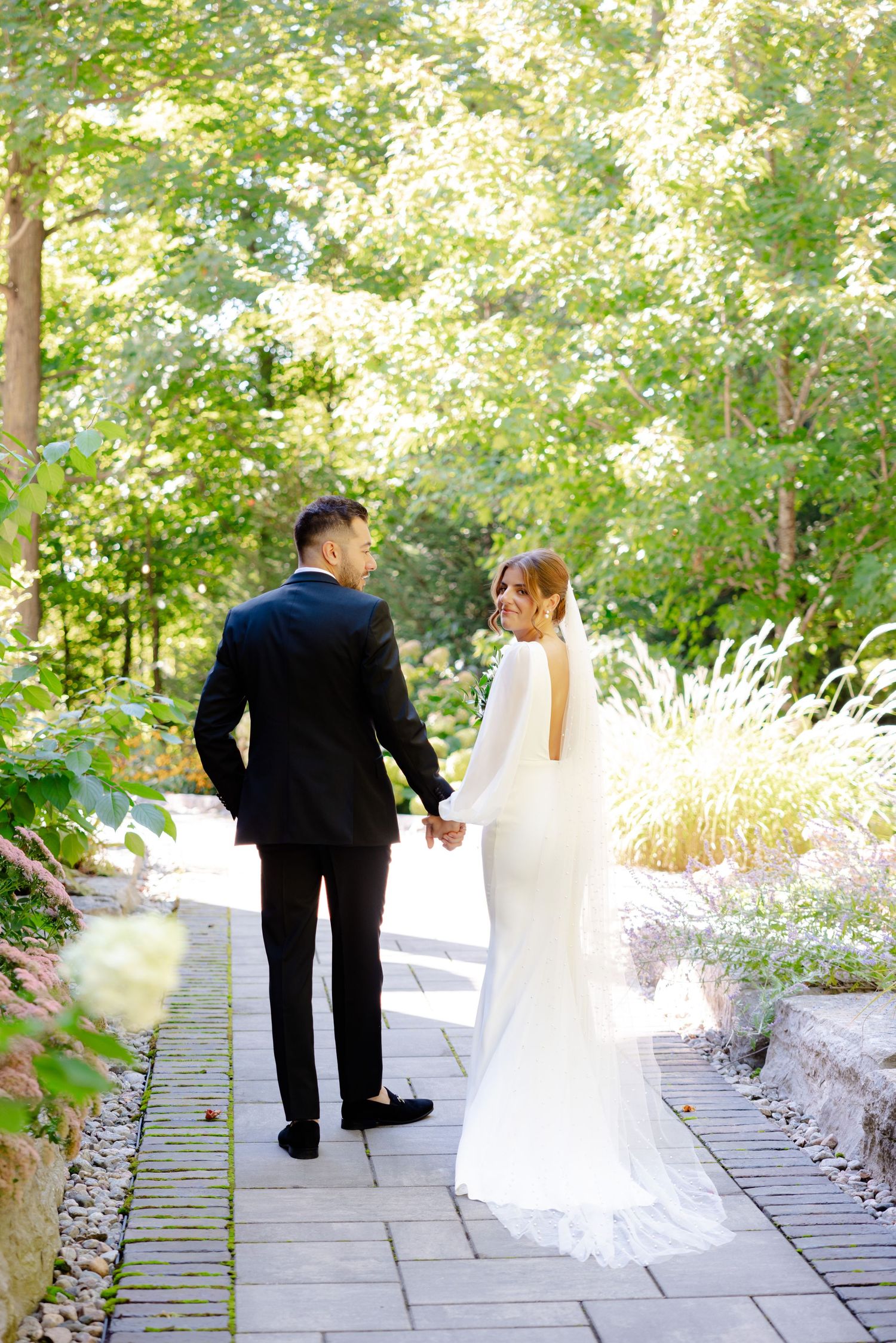 A bride and groom hold hands while walking down a landscaped stone path surrounded by summer foliage.