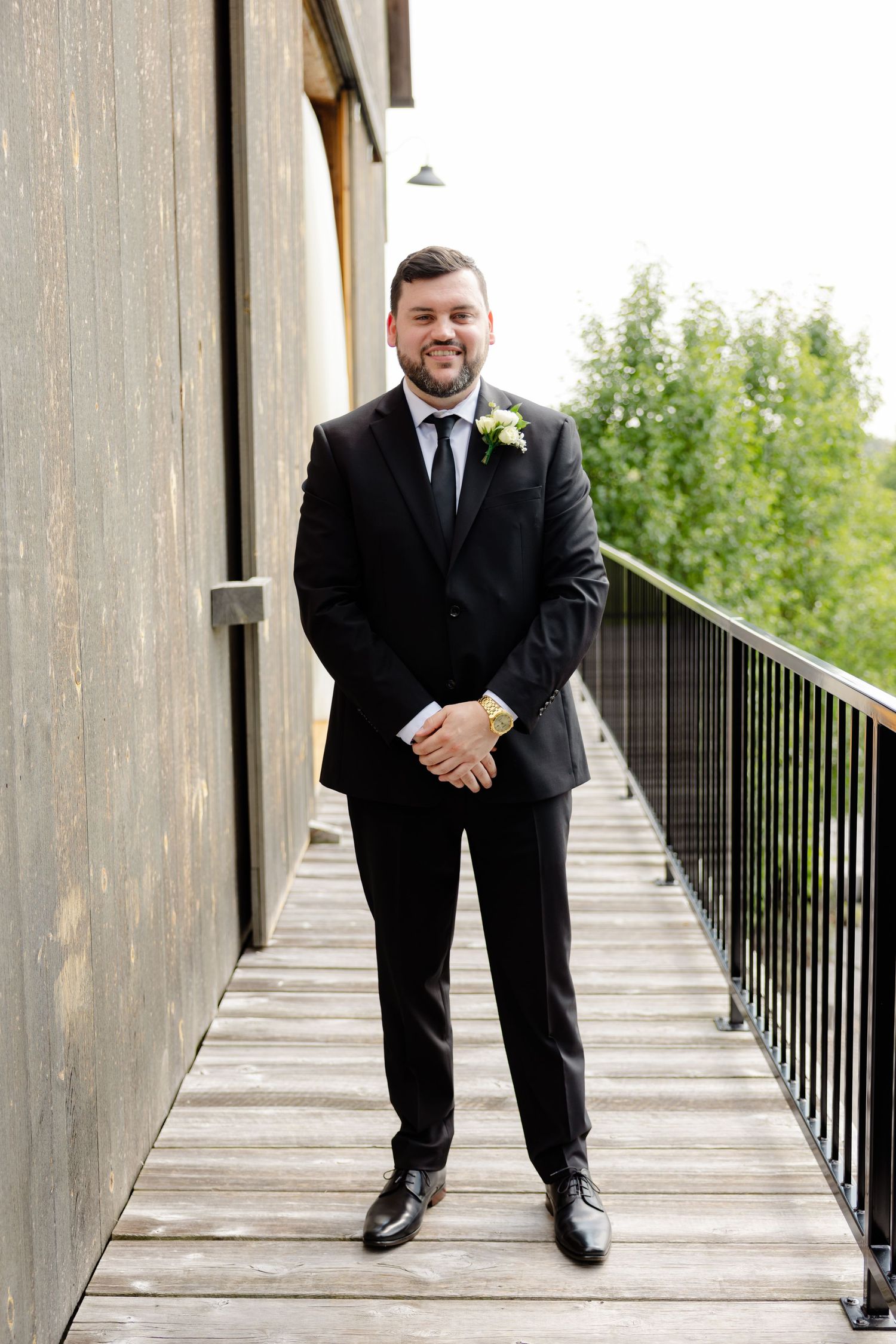 Person in black suit with boutonniere stands on wooden deck during formal event.