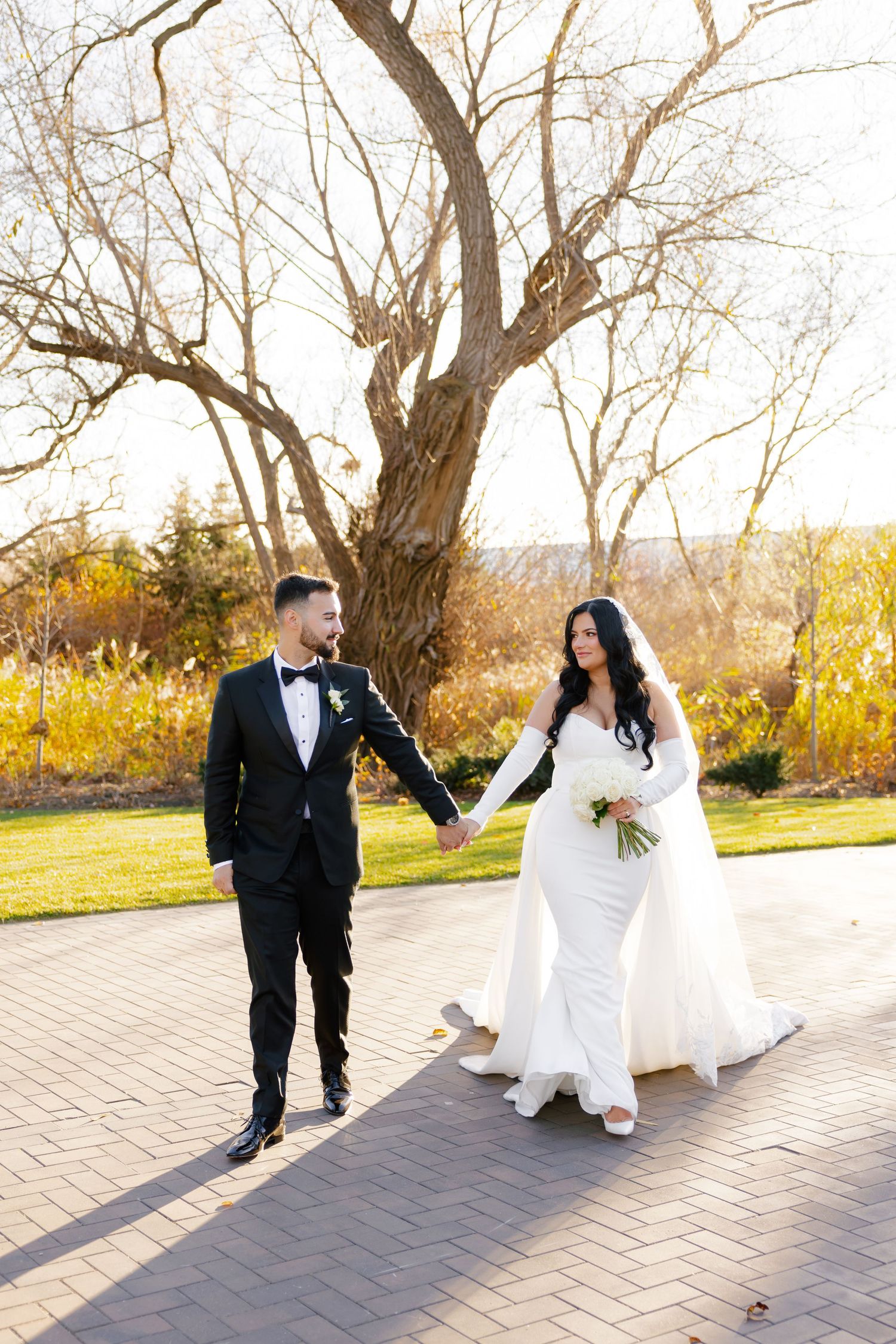 Newlyweds walking hand in hand on brick pathway with bare trees and fall foliage.