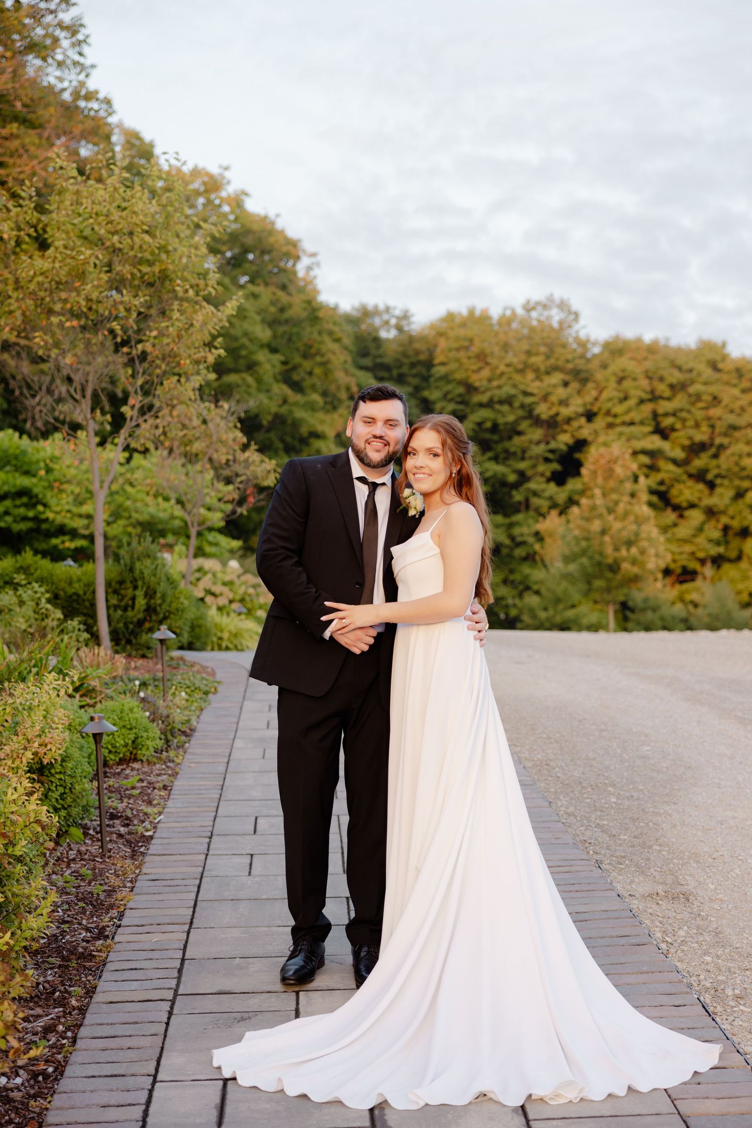 Wedding couple poses on stone walkway surrounded by autumn trees and landscaping.
