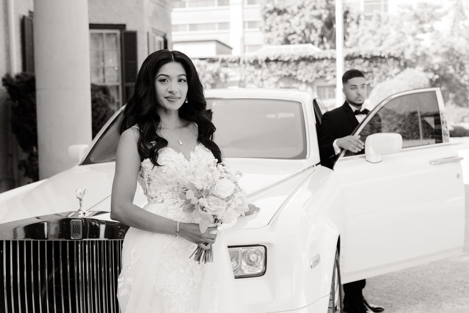 A bride in a white lace dress poses beside a classic Rolls Royce luxury car.