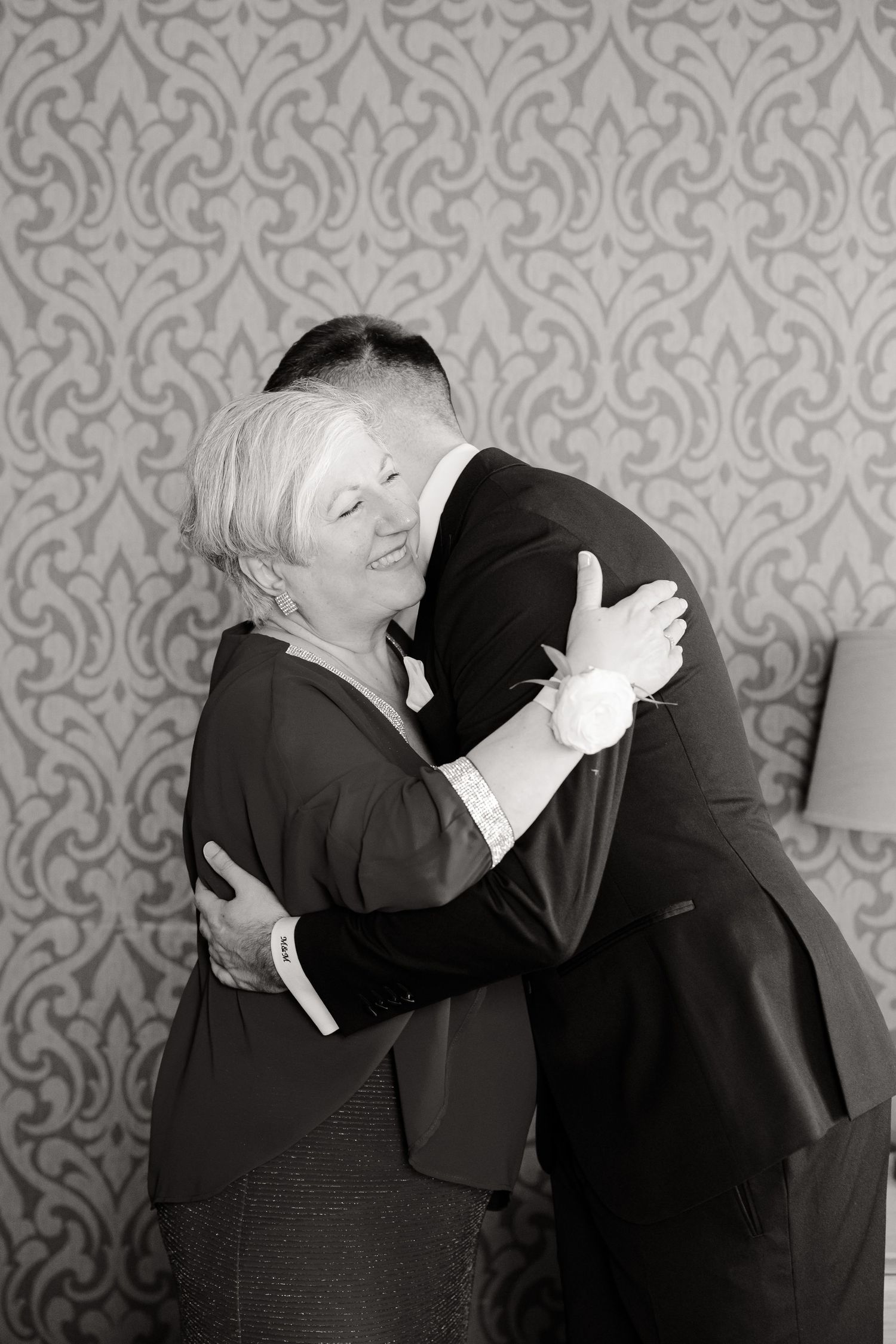 A heartwarming embrace between a woman in black and a person in formal attire against a patterned wallpaper.