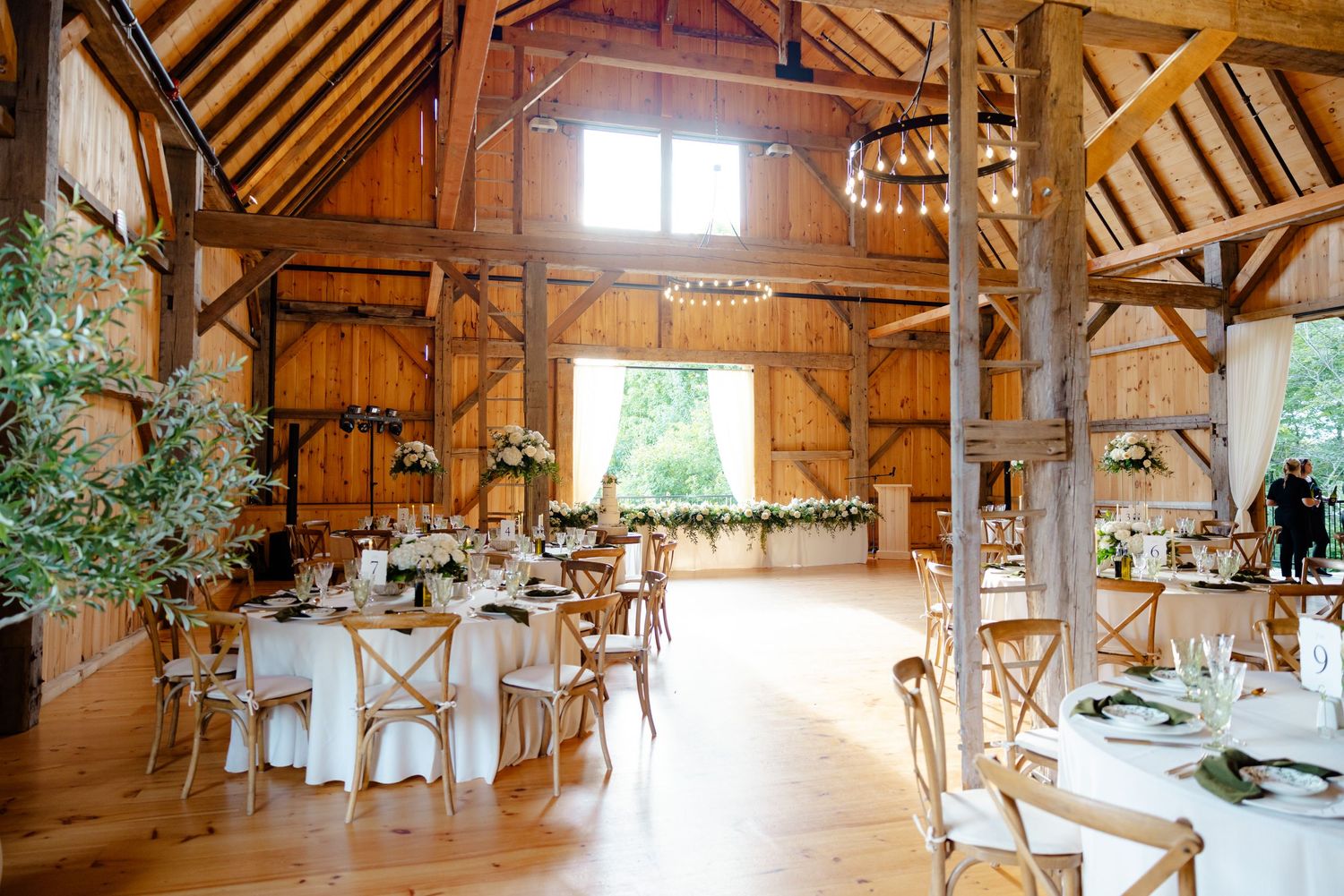 A bright rustic barn wedding venue with wooden beams, exposed rafters, and round dining tables.