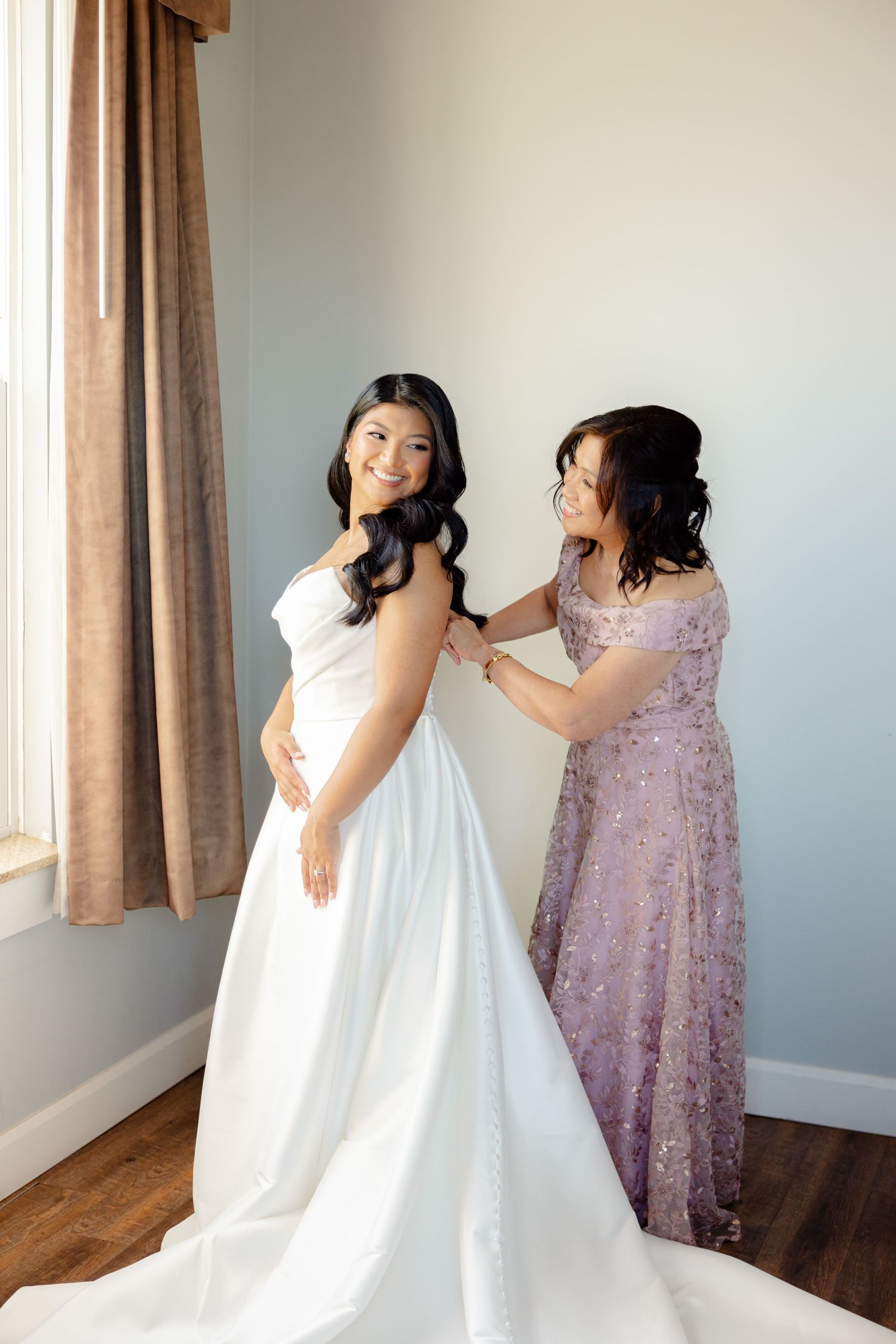 Two people share a joyful moment while preparing in a simple room with brown curtains and hardwood floors.