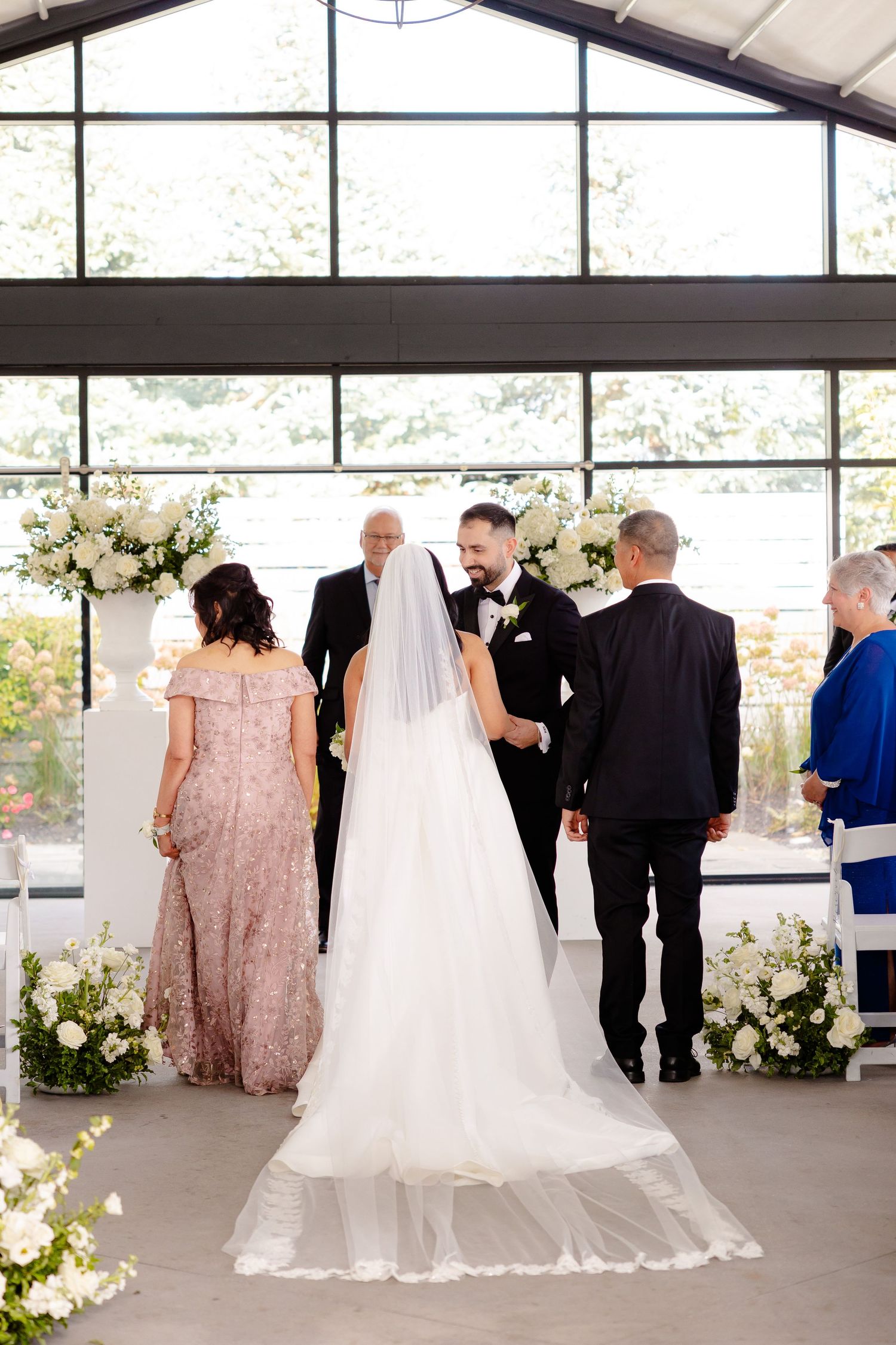 Wedding ceremony in a bright glass venue with floral arrangements and guests gathered around the couple at the altar.