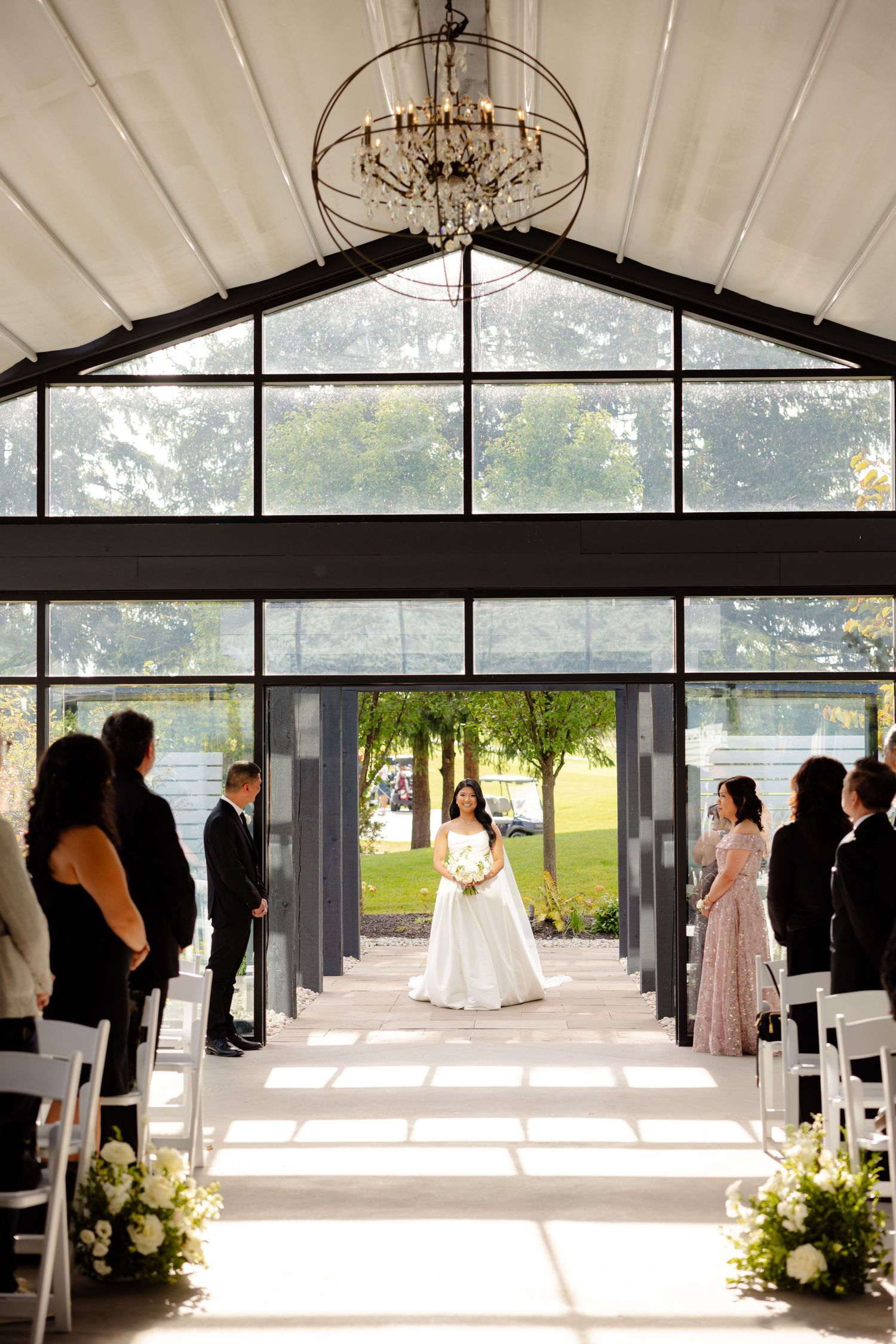 Wedding ceremony setup in a modern glass venue with chandelier and white floral aisle decorations.