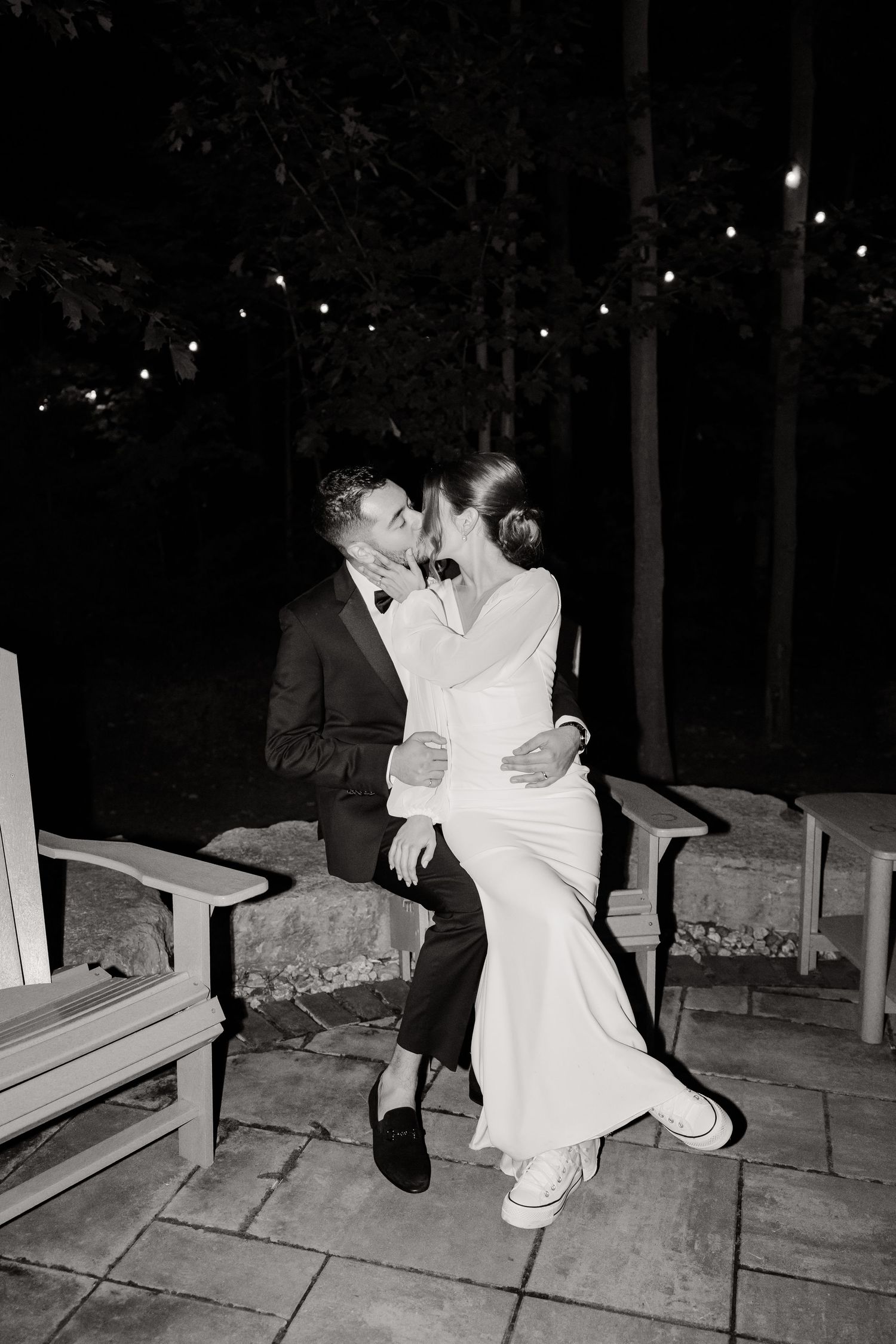Black and white photo of newlyweds sharing romantic moment on outdoor bench at night.
