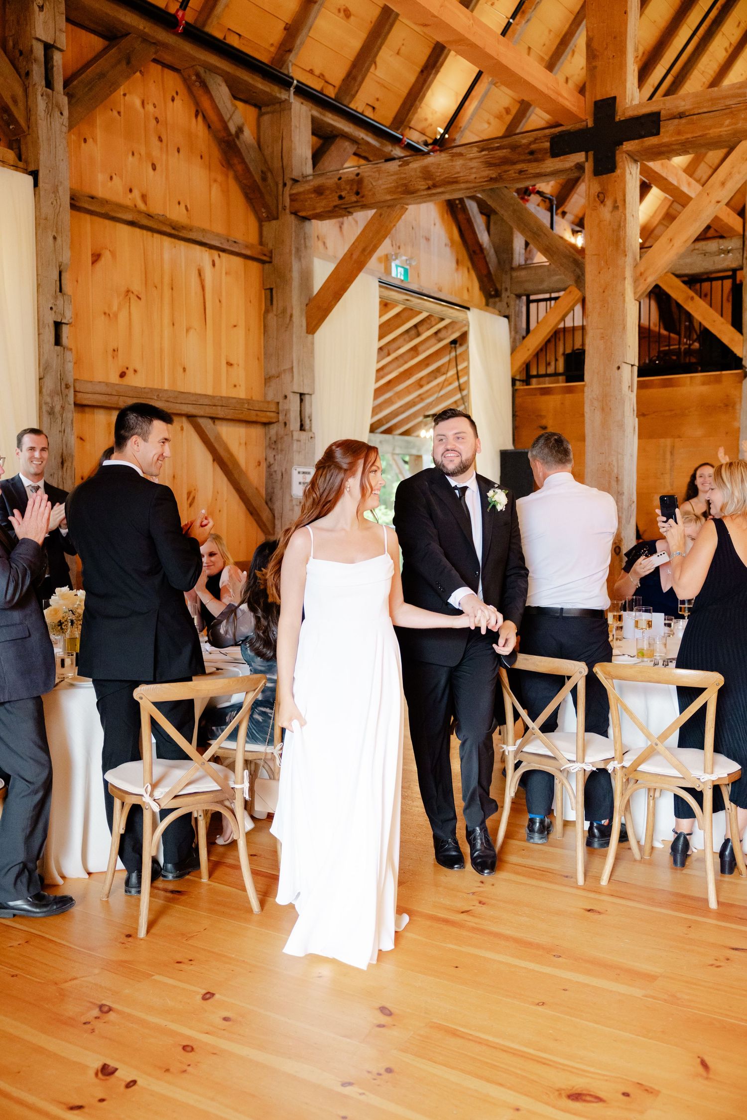 Guests applaud during a wedding celebration inside a timber-framed barn with natural wood floors.