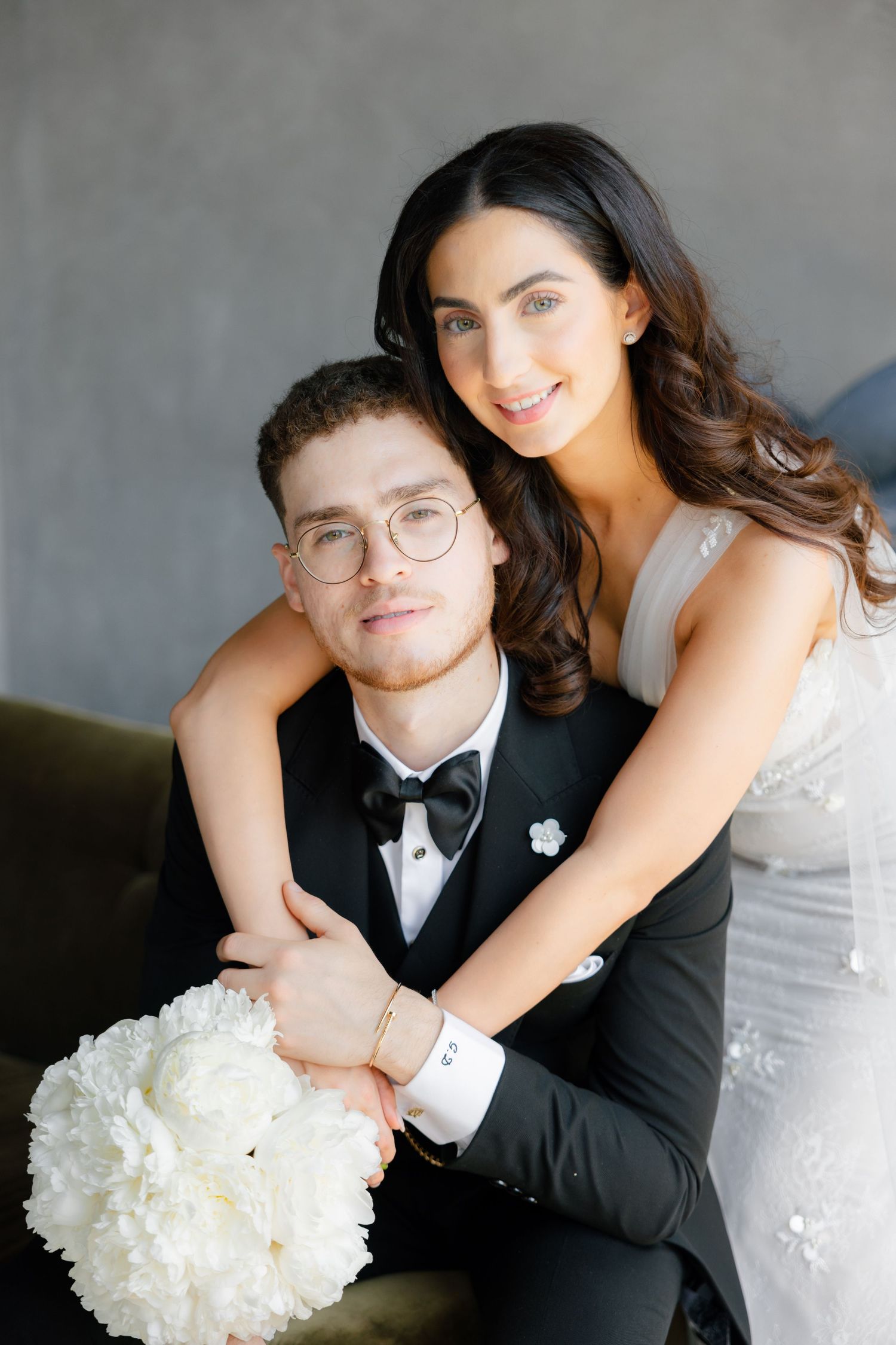 Joyful wedding portrait of couple embracing on couch with white floral bouquet.