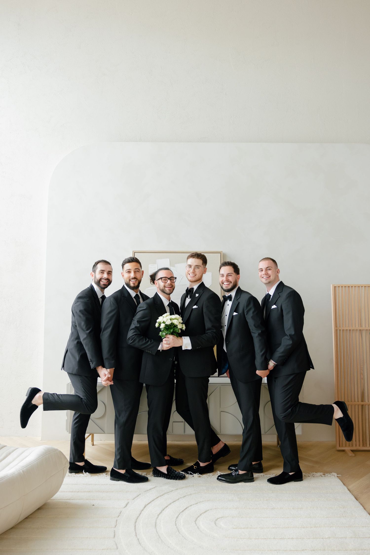 Groomsmen in black suits striking playful pose against white wall.